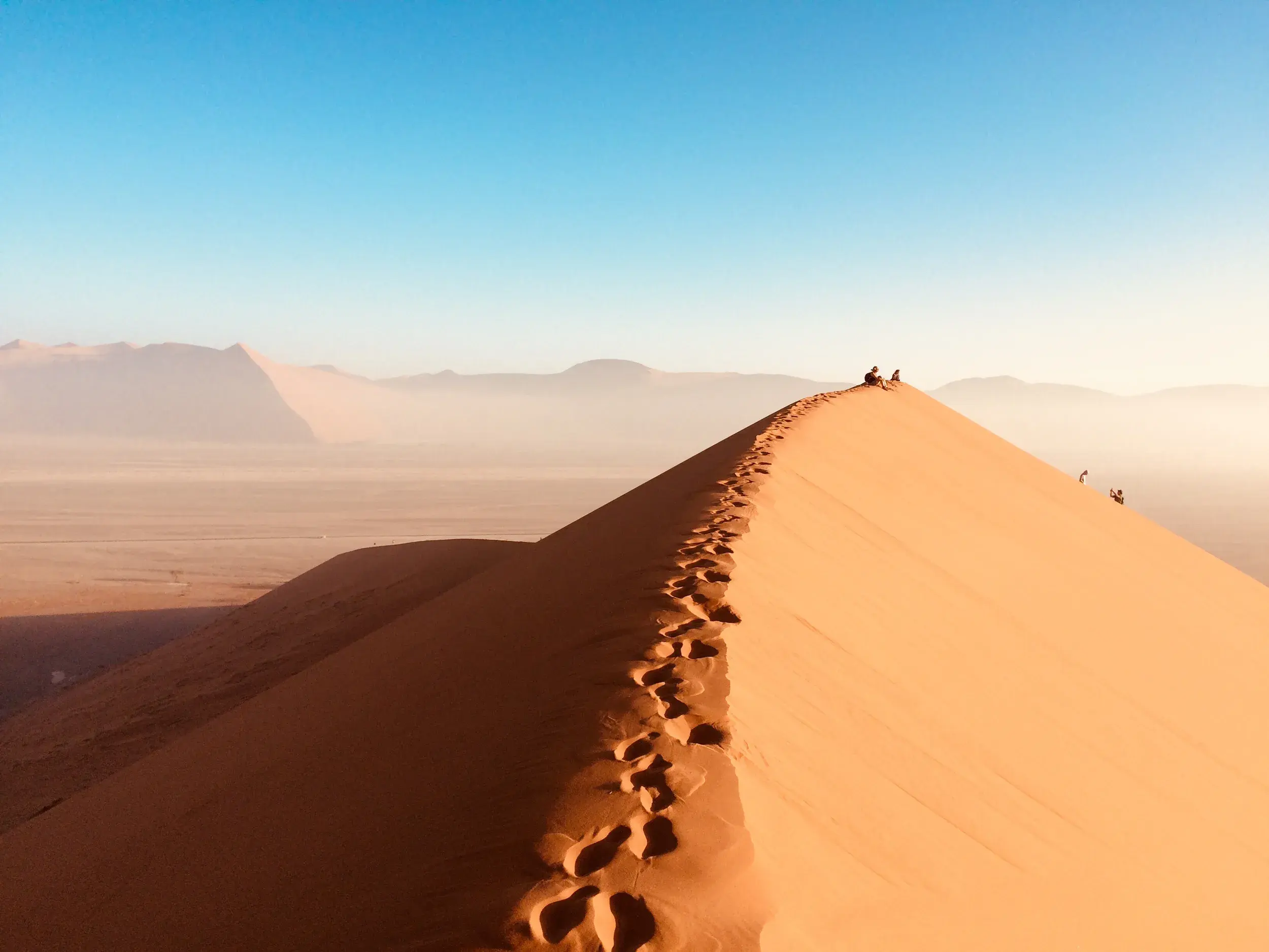 Eine kleine Gruppe von Reisenden sitzt auf dem schmalen Grat einer hohen, orangen Sanddüne im Sossusvlei bei Sonnenaufgang.