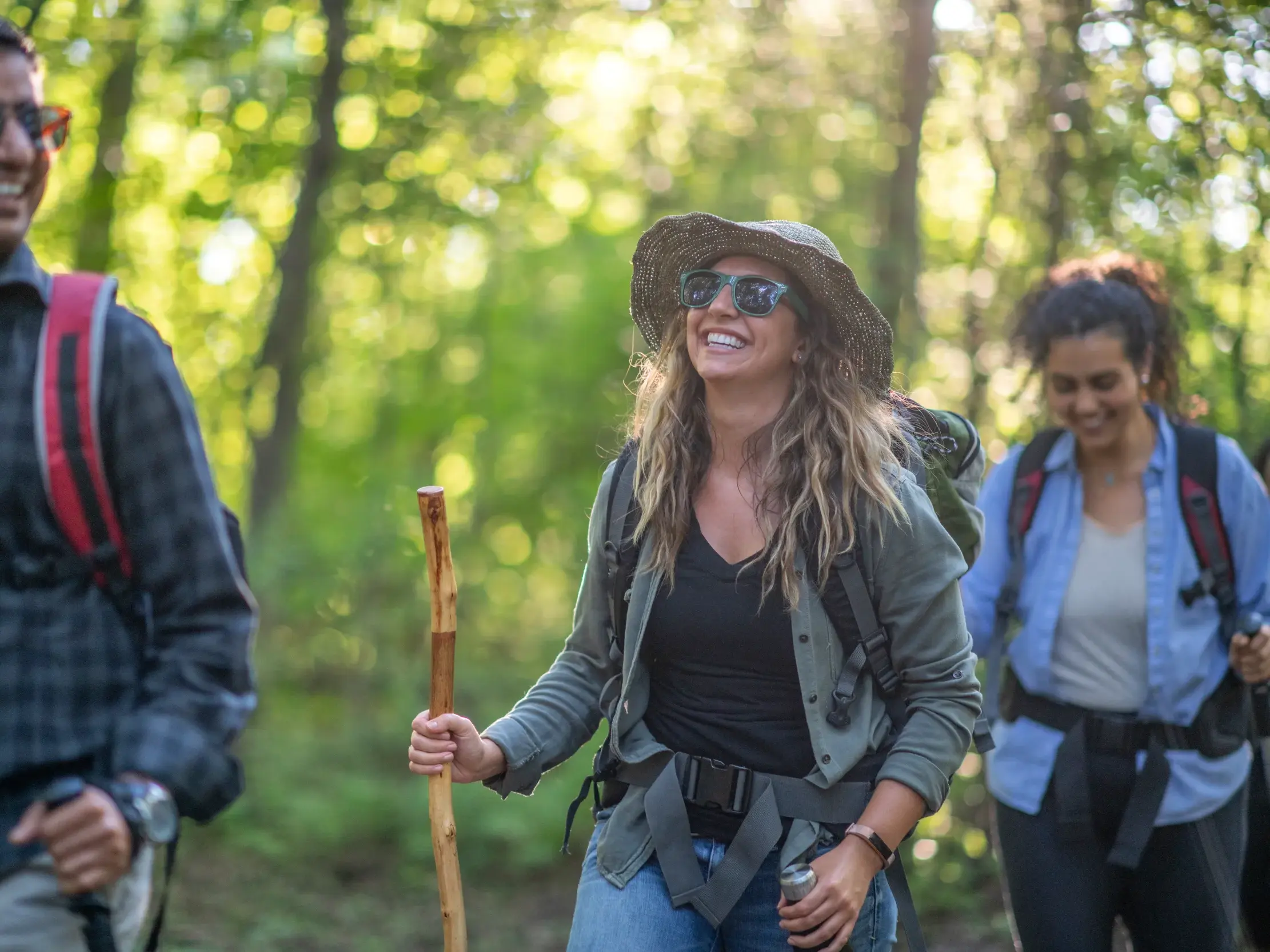 Eine junge Frau lacht bei einer Gruppenreise in Ecuador, während sie mit Wanderstock und Rucksack durch einen Wald wandert.