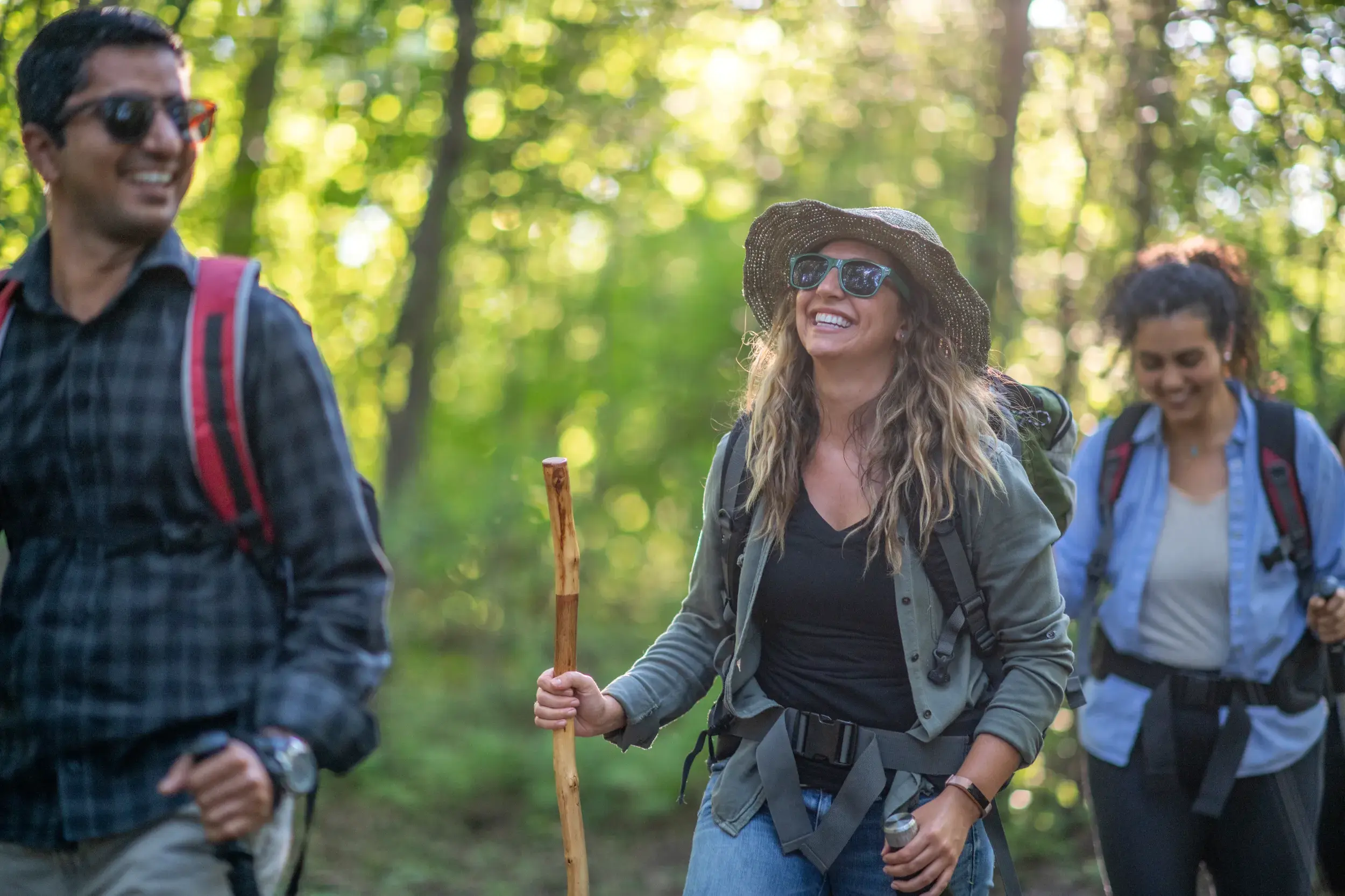 Eine junge Frau lacht bei einer Gruppenreise in Ecuador, während sie mit Wanderstock und Rucksack durch einen Wald wandert.