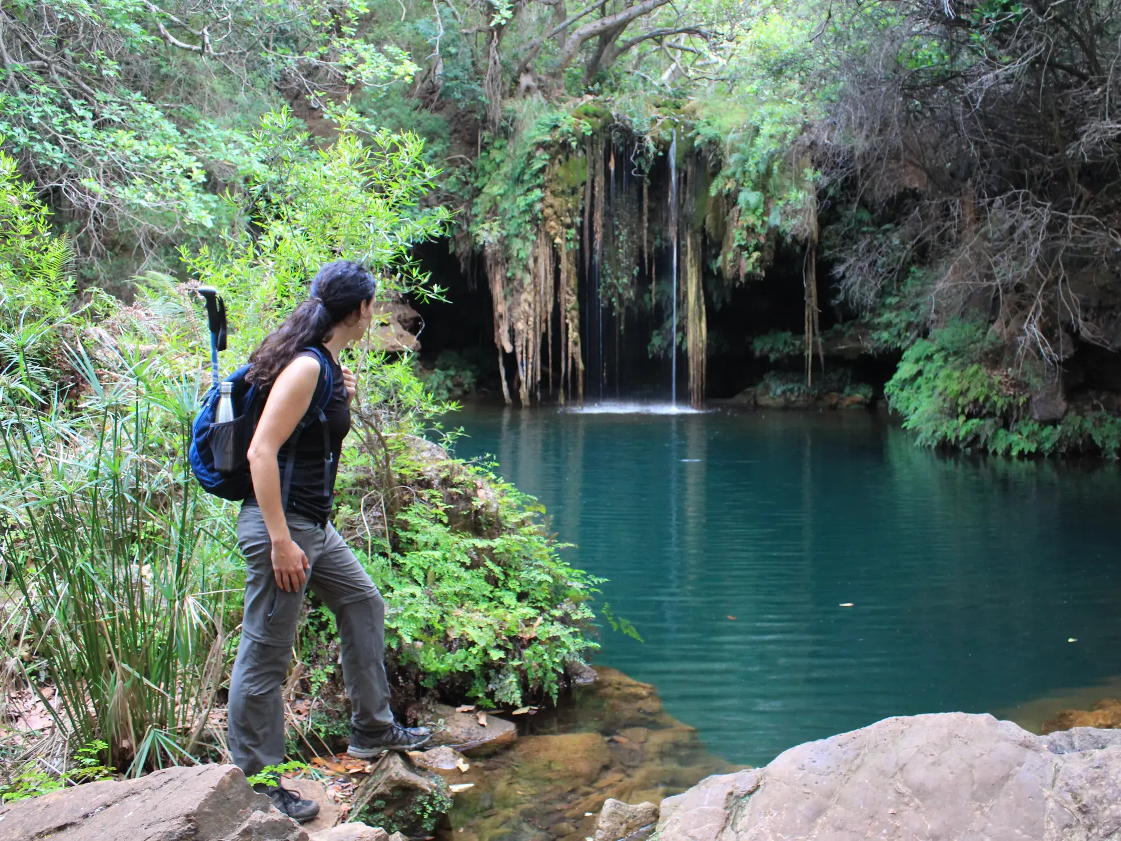 Eine Wanderin mit Rucksack steht am Rande eines türkisen Naturpools in Südafrika und blickt auf einen von Pflanzen umrankten Wasserfall.