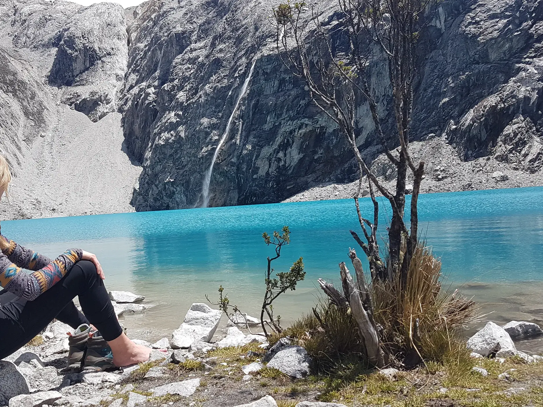 Eine Wanderin ruht sich barfuß am Ufer der leuchtend blauen Laguna 69 in Peru vor einer Felswand mit Wasserfall aus.