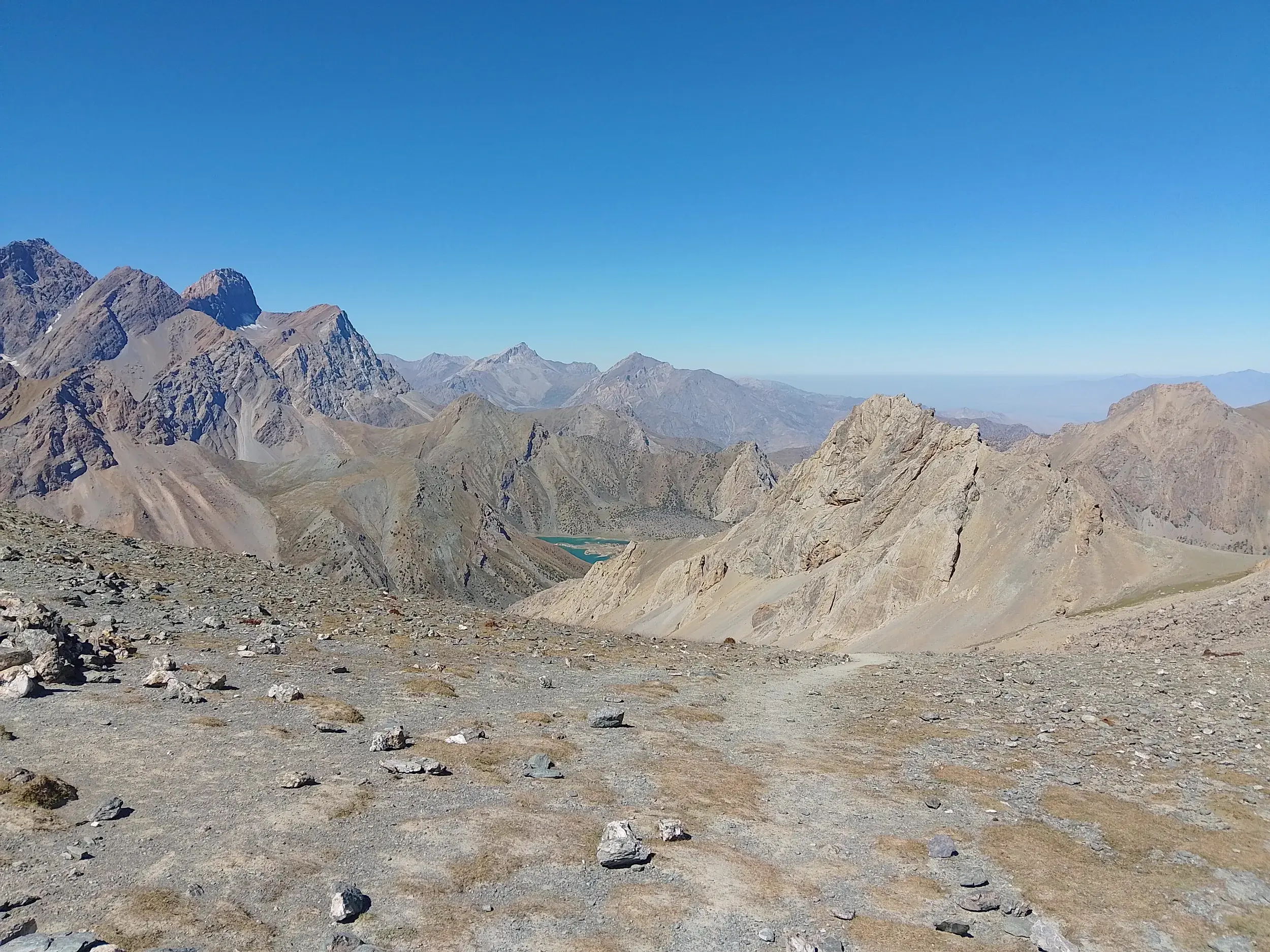 Weitblick von einem steinigen Pass auf die schroffen Gipfel und einen türkisen Bergsee während einer Trekking-Reise im Fan-Gebirge.