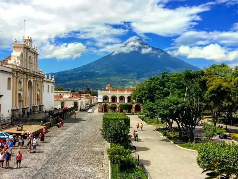 Lebhafter Platz in Antigua Guatemala mit Kolonialbauten, grünen Bäumen und dem imposanten Vulkan Agua im Hintergrund.
