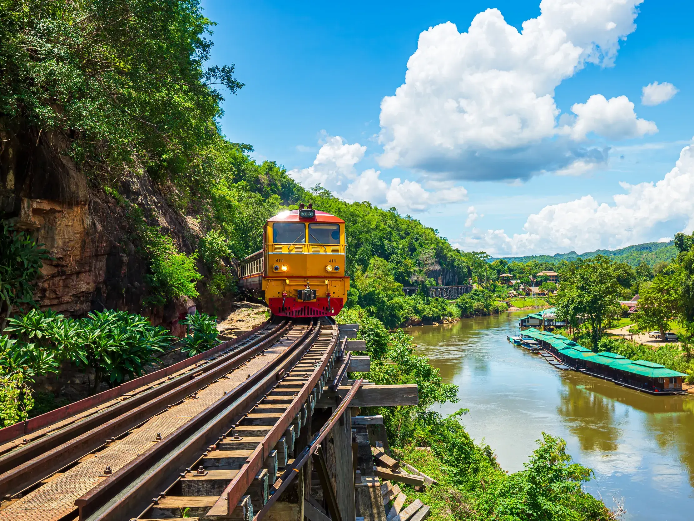 Ein gelber Zug fährt auf der malerischen Strecke der Death Railway am River Kwai entlang einer steilen Felswand.
