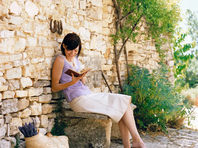 Eine lächelnde Frau liest, an eine rustikale Mauer gelehnt, auf einer Steinbank in der Provence, neben ihr ein Korb Lavendel.
