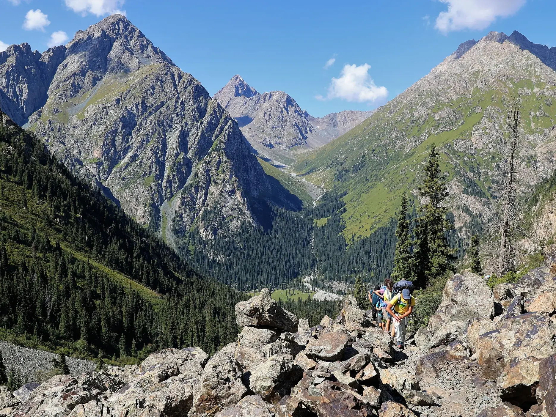 Trekking-Gruppe steigt über Felsen im Terskej-Alatau-Gebirge in Kirgistan mit Panoramablick auf ein tiefes, grünes Tal.