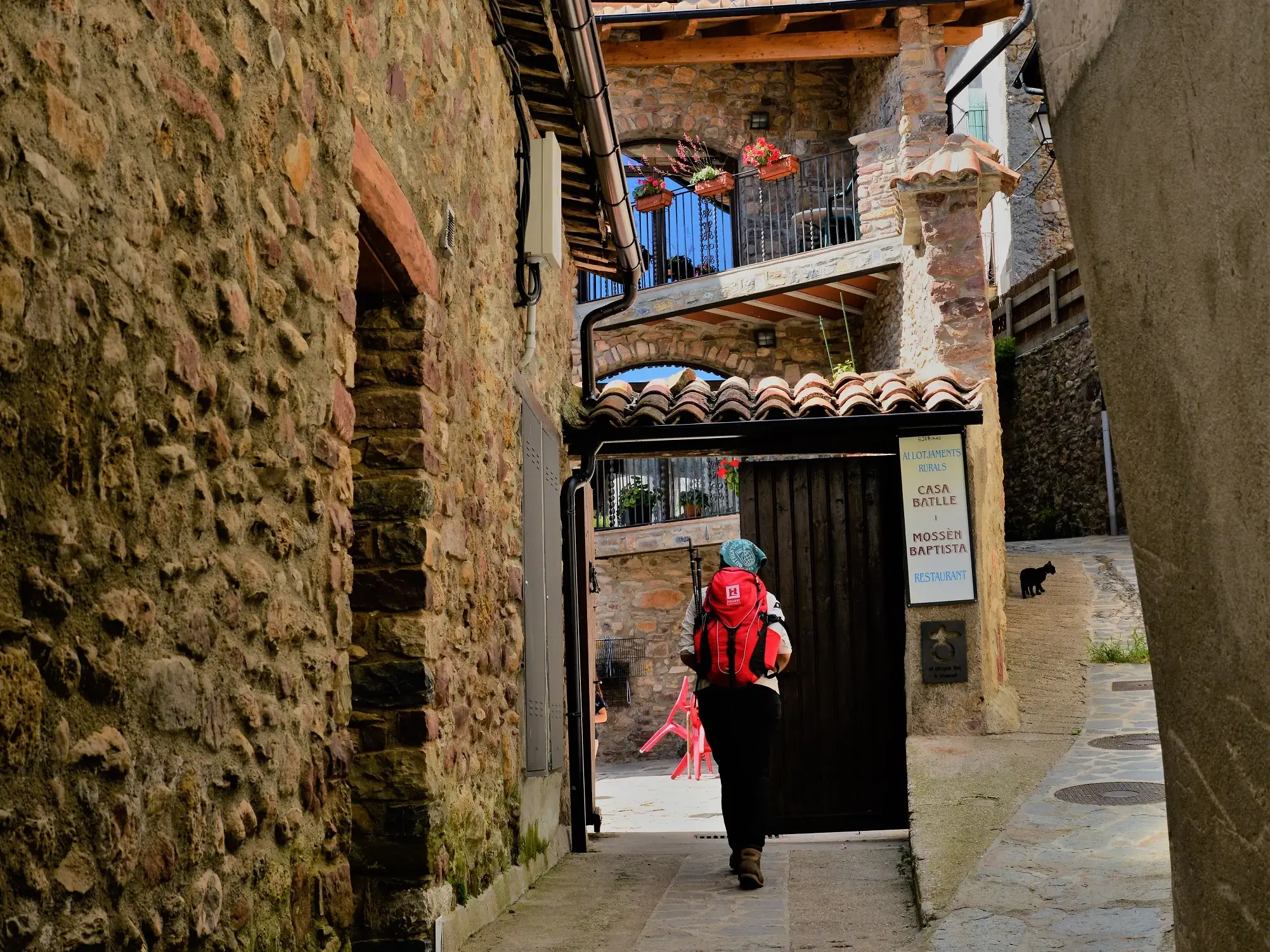 Wanderer mit rotem Rucksack beim Trekking in einer engen Gasse mit Steinhäusern in einem authentischen Dorf der Pyrenäen.
