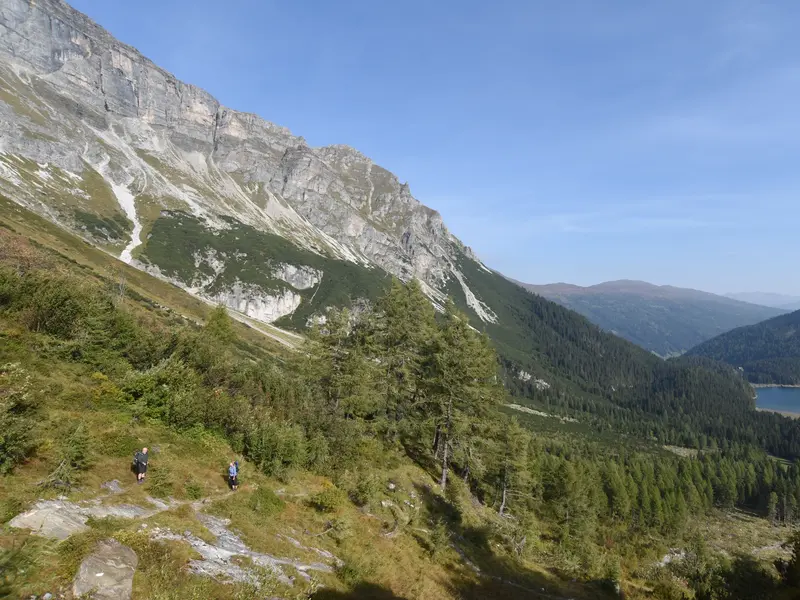 Zwei Wanderer auf einem schmalen Pfad am Hang mit weitem Blick über das Tal auf den Obernberger See in den Stubaier Alpen.