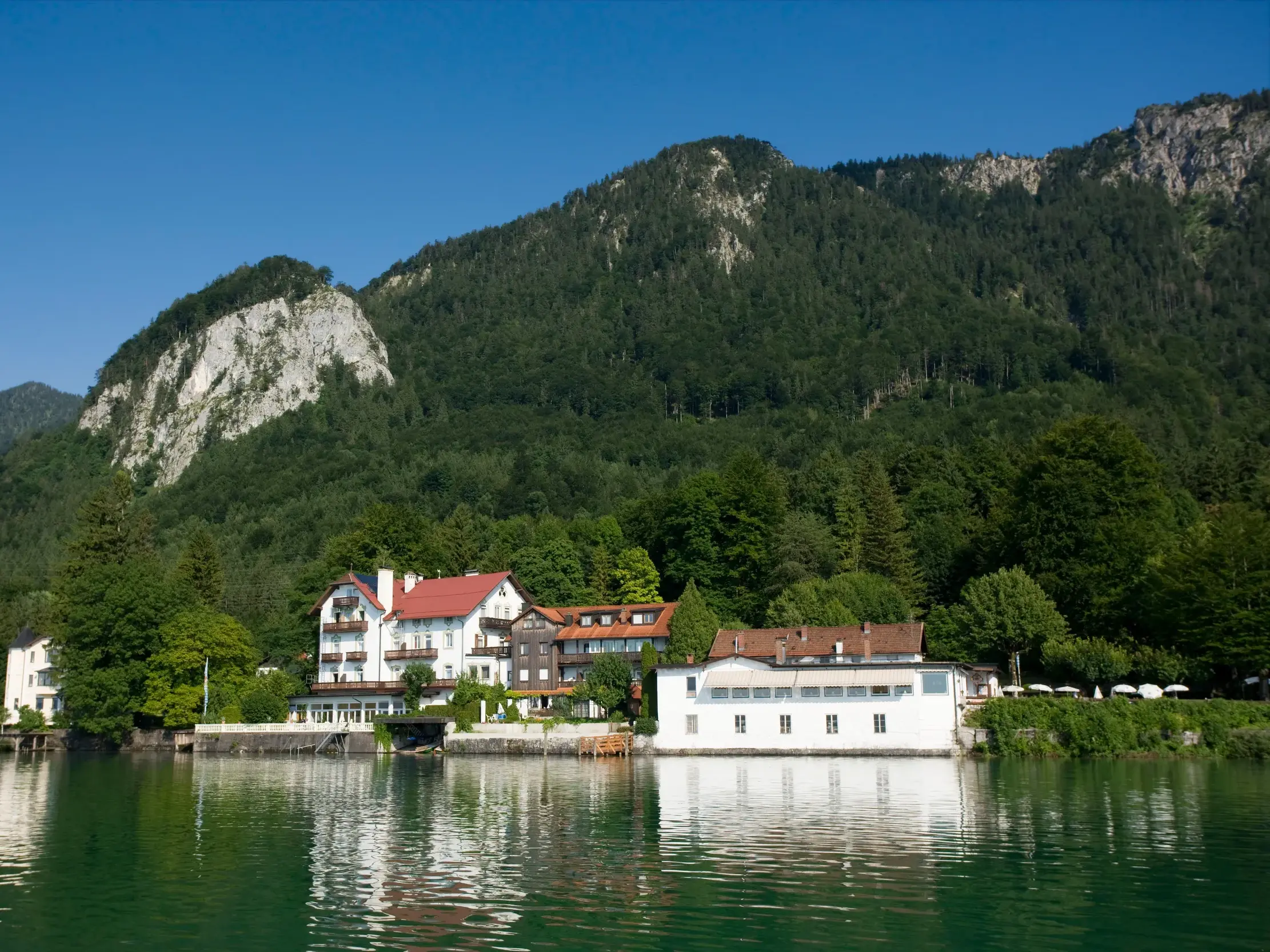 Das Seehotel Grauer Bär am Kochelsee, Ausgangspunkt für Wanderungen, spiegelt sich im Wasser vor alpiner Bergkulisse.