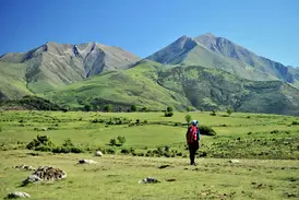 Trekking in den spanischen Pyrenäen: Eine Person mit Rucksack blickt über grüne, hügelige Weiden auf eine Bergkette.