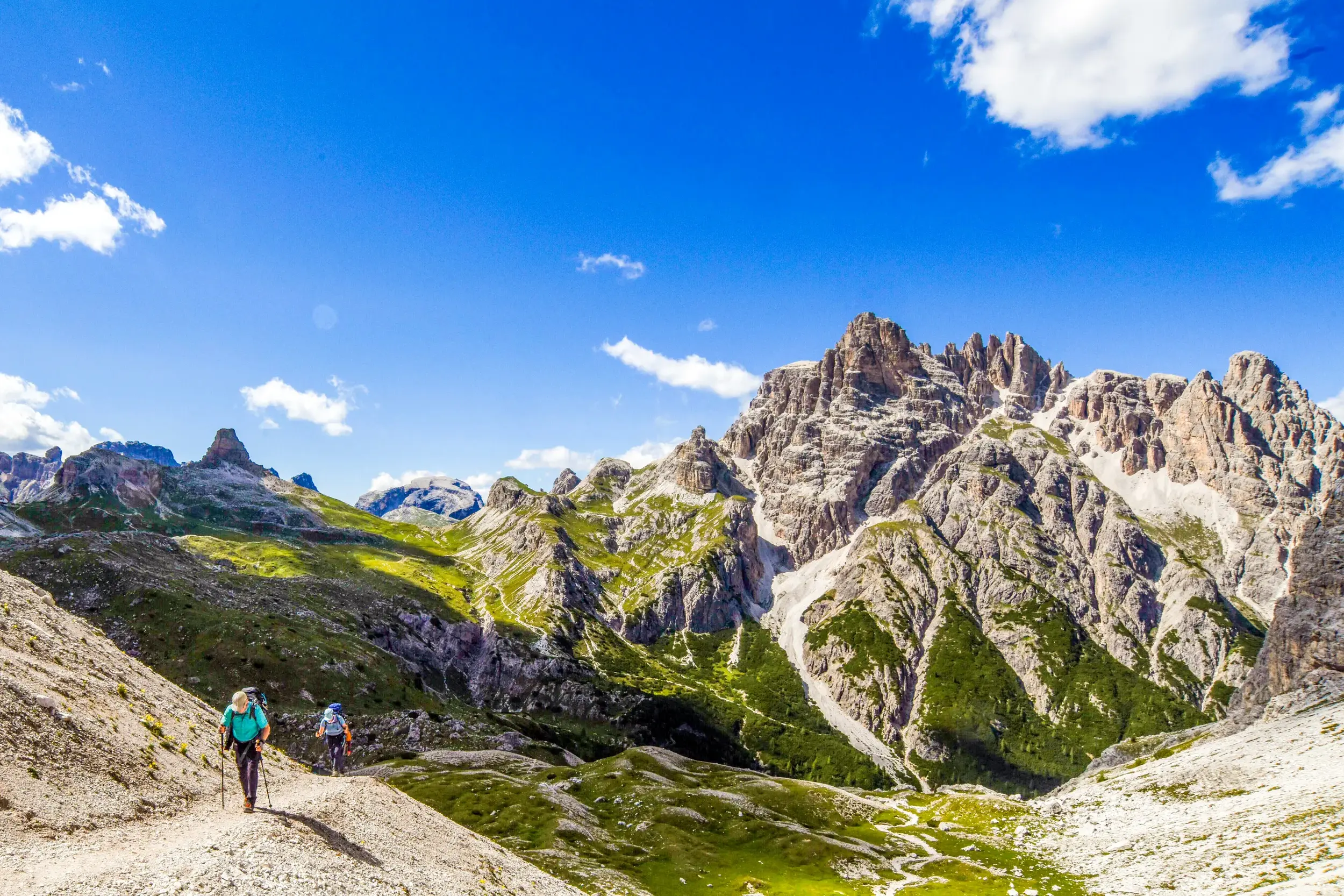 Zwei Wanderer mit Stöcken auf einer Trekking-Tour vor der Felskulisse der Sextener Dolomiten bei strahlendem Sonnenschein.