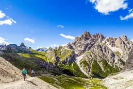 Zwei Wanderer mit Stöcken auf einer Trekking-Tour vor der Felskulisse der Sextener Dolomiten bei strahlendem Sonnenschein.