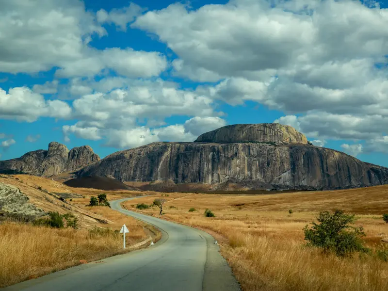 Kurvige Landstraße durch eine Savannenlandschaft zum imposanten Granitfelsen des Andringitra-Gebirges in Madagaskar.