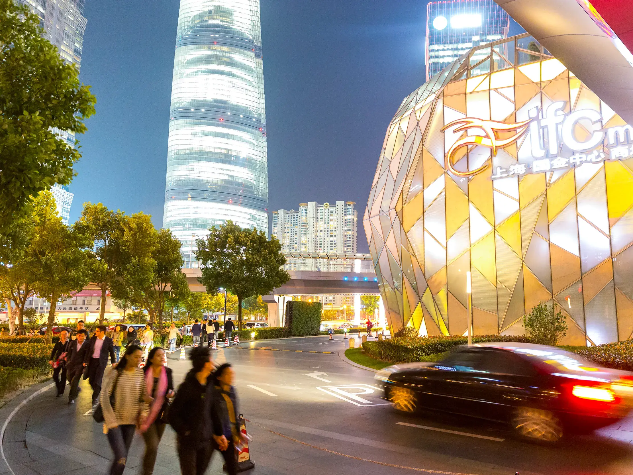Passanten spazieren am Abend vor der leuchtenden Fassade der IFC Mall und dem Shanghai Tower im modernen Stadtteil Pudong.