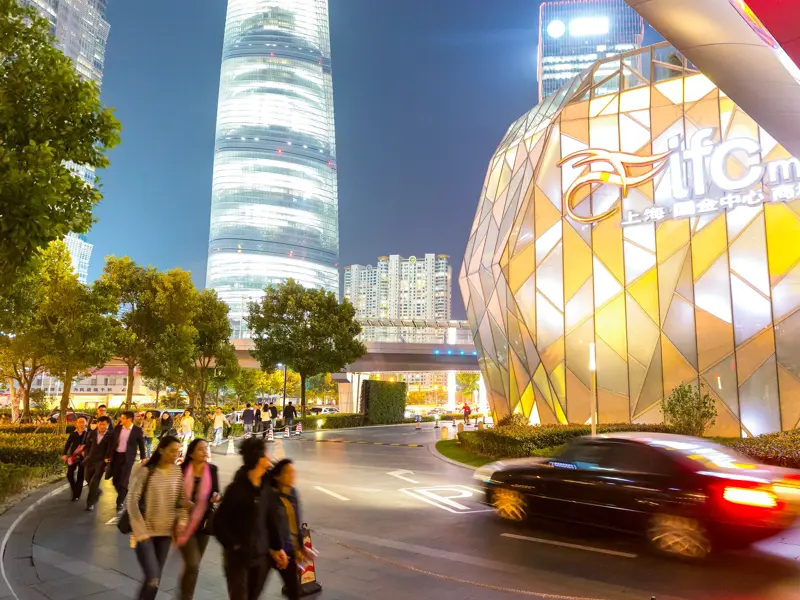 Passanten spazieren am Abend vor der leuchtenden Fassade der IFC Mall und dem Shanghai Tower im modernen Stadtteil Pudong.