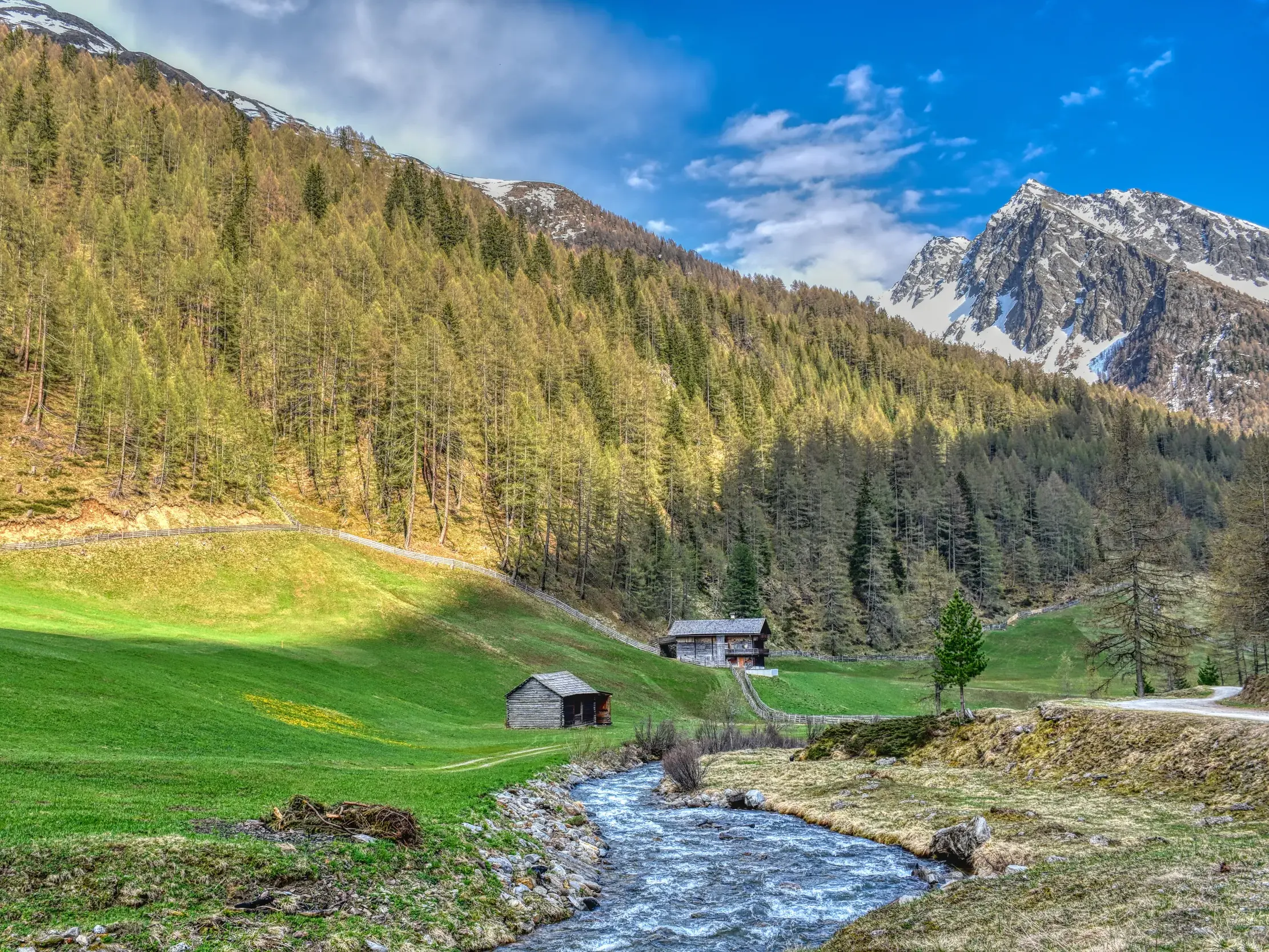 Ein Bergbach im Villgratental, flankiert von grünen Wiesen mit Almhütten und einem schneebedeckten Gipfel in Osttirol.