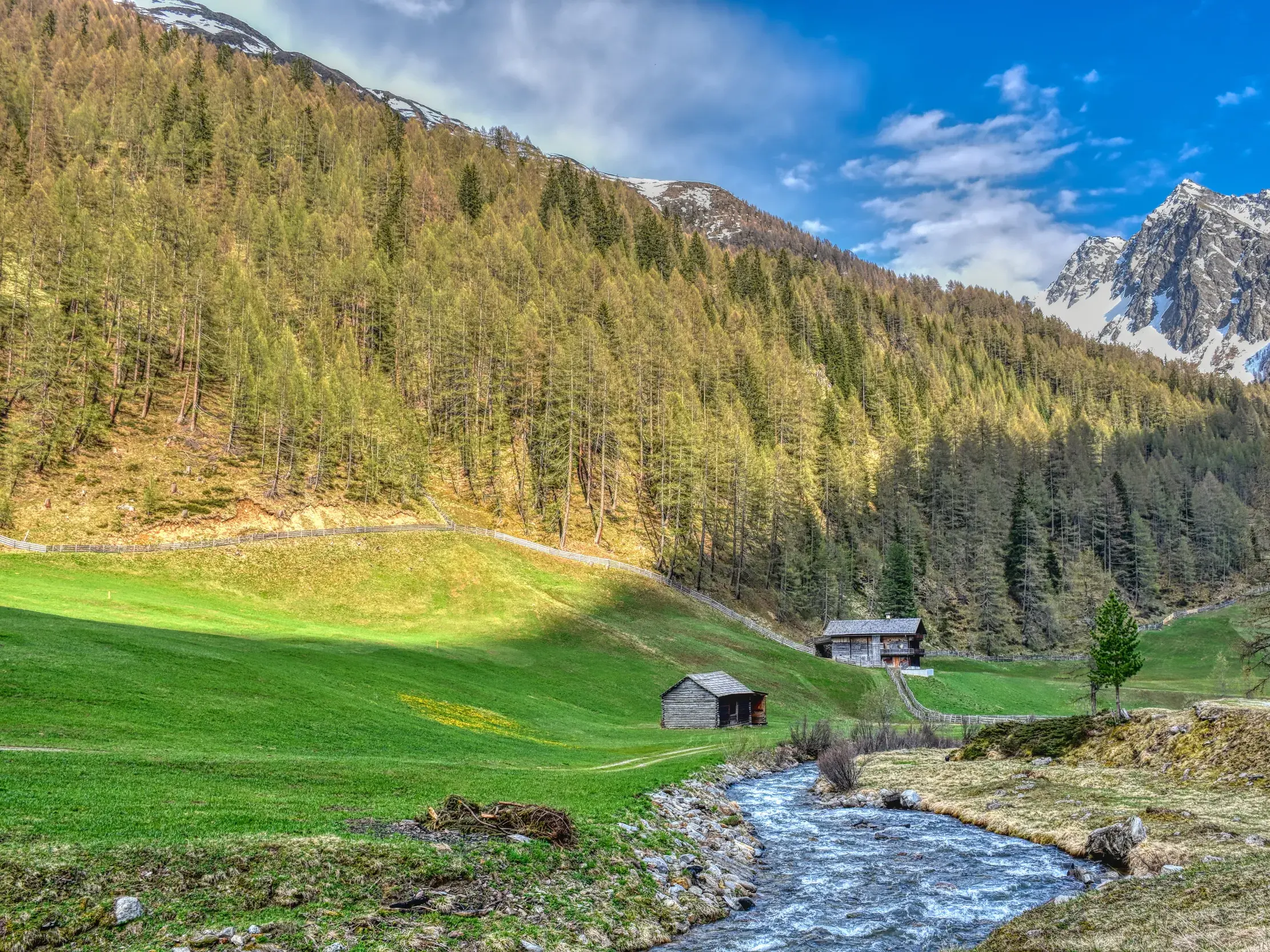 Rauschender Bergbach im idyllischen Winkeltal in Osttirol mit Almhütten vor der Kulisse schneebedeckter Alpengipfel.