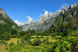 Sonniger Blick in ein einsames Tal in Albanien mit grünen Wiesen und schroffen, zackigen Berggipfeln im Hintergrund.