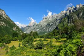 Sonniger Blick in ein einsames Tal in Albanien mit grünen Wiesen und schroffen, zackigen Berggipfeln im Hintergrund.