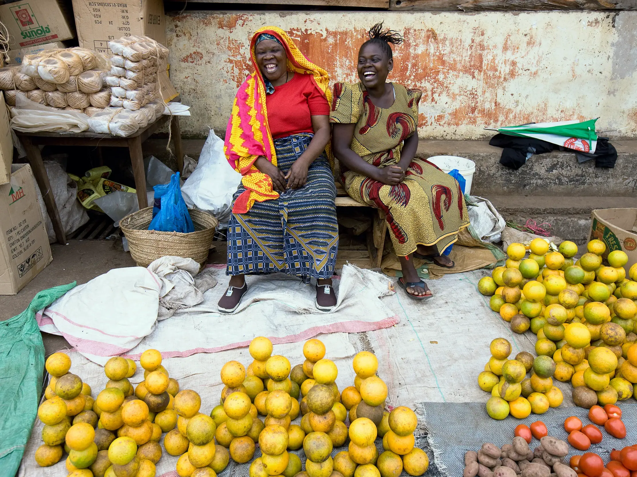 Zwei Händlerinnen in bunter, traditioneller Kleidung lachen herzlich an ihrem Marktstand in Lushoto mit Orangen und Gemüse.