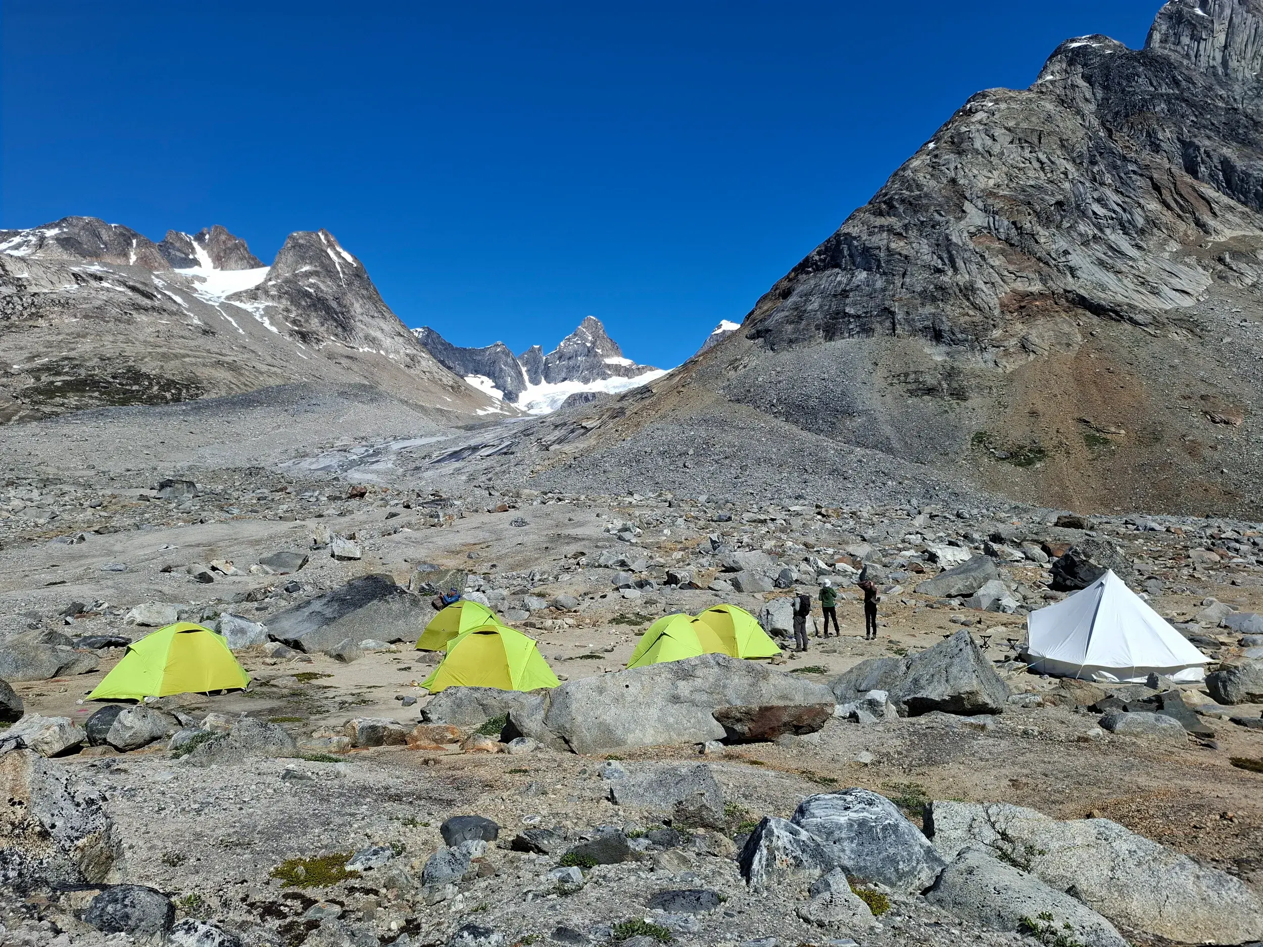 Eine kleine Reisegruppe steht im Zeltcamp in der arktischen Felslandschaft Ostgrönlands mit Blick auf die Niialigaq-Gipfel.