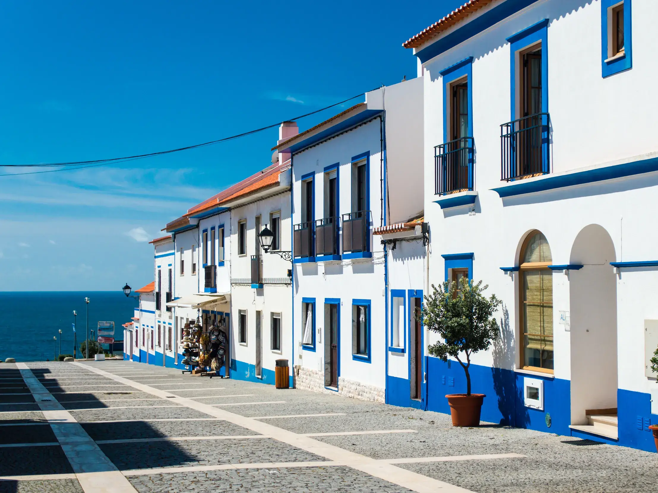 Eine gepflasterte Straße in Porto Covo führt entlang weiß-blauer Häuserfassaden hinunter zum blauen Atlantik.