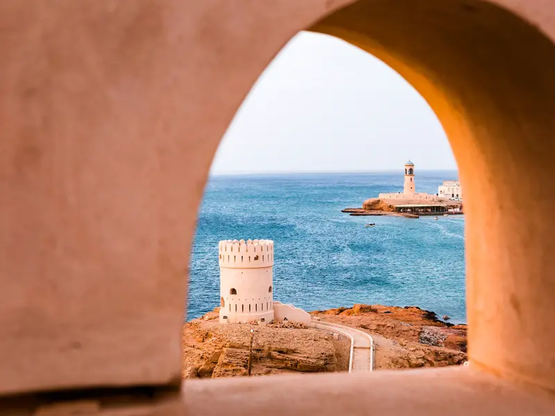 Gerahmter Ausblick durch ein Lehmfenster auf den historischen Wachturm von Sur und das türkisblaue Meer des Omans.