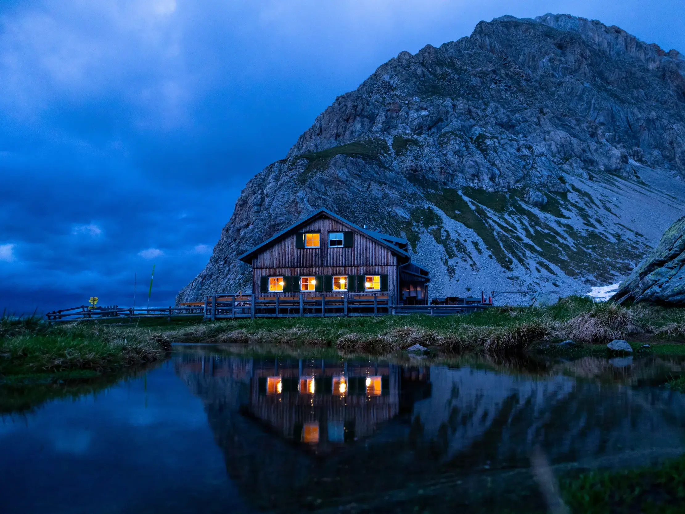 Die Wolayerseehütte am Fuße der Hohen Warte spiegelt sich mit ihren erleuchteten Fenstern im Wolayersee bei Dämmerung.