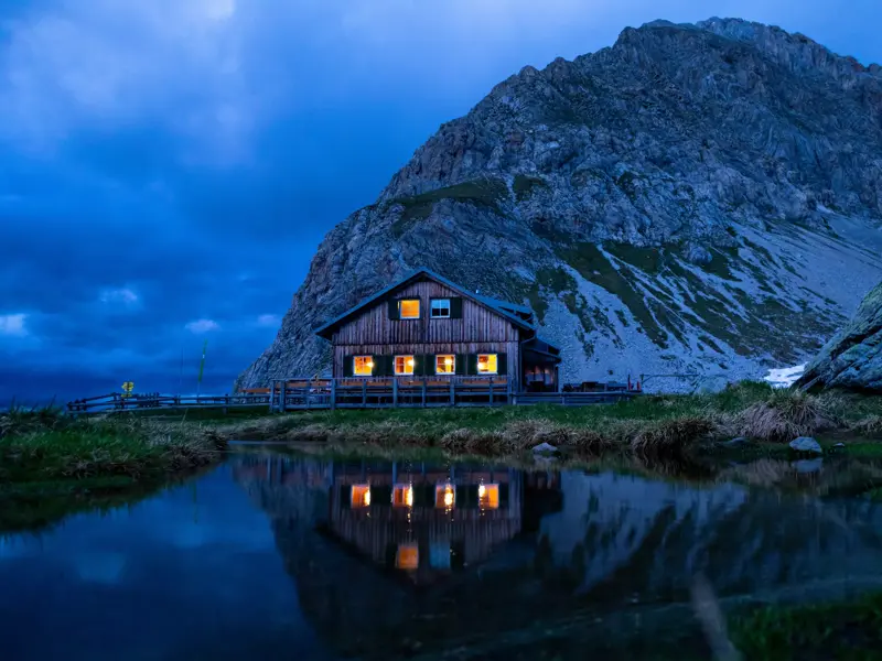 Die beleuchtete Wolayerseehütte am Karnischen Höhenweg spiegelt sich bei Dämmerung im ruhigen Wasser des Wolayer Sees.