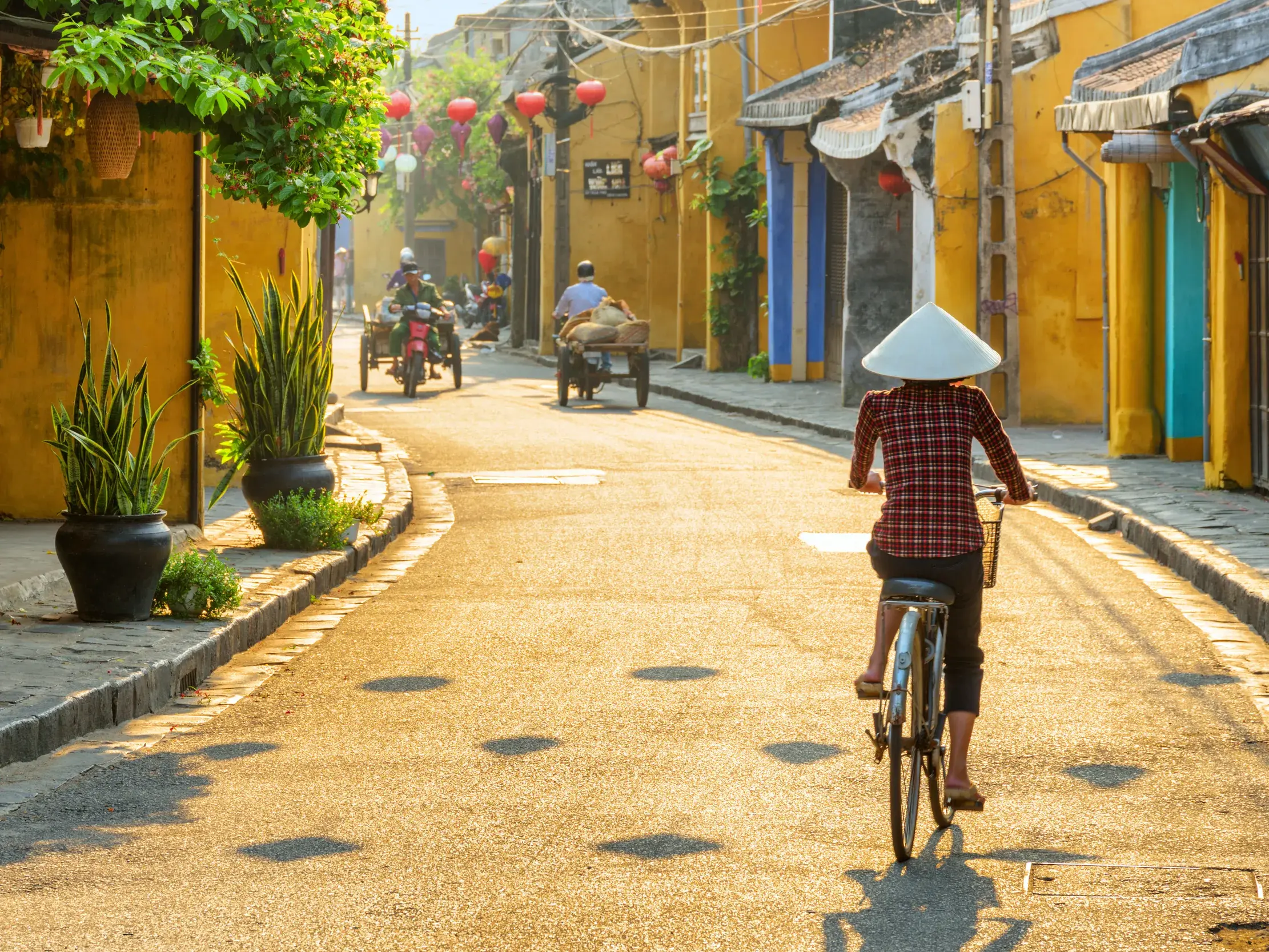 Eine Person mit traditionellem Kegelhut radelt durch eine sonnige Gasse der Altstadt von Hoi An mit gelben Häusern.