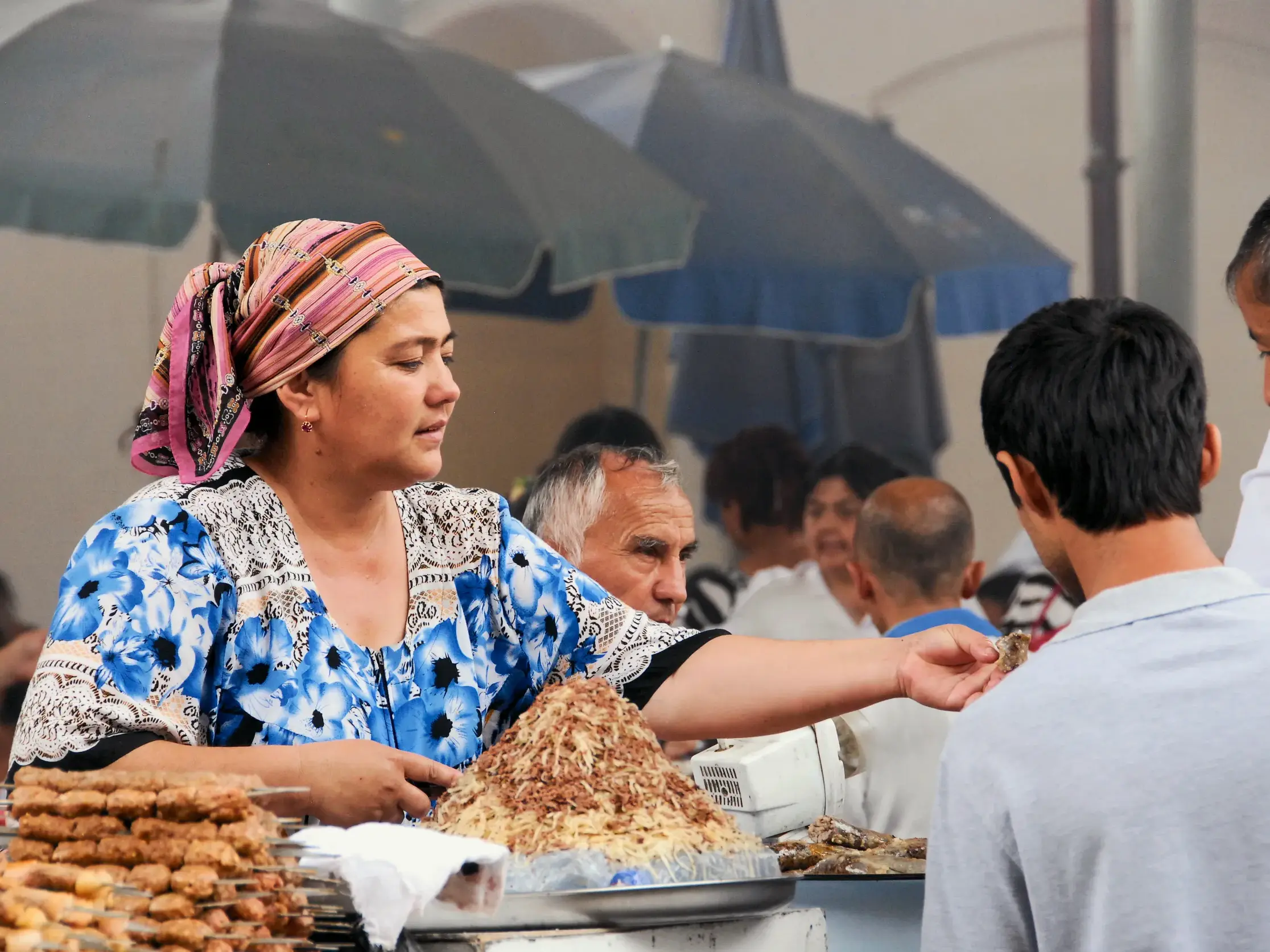 Eine usbekische Händlerin mit buntem Kopftuch bietet auf einem Basar einem Kunden eine Kostprobe orientalischer Spezialitäten an.