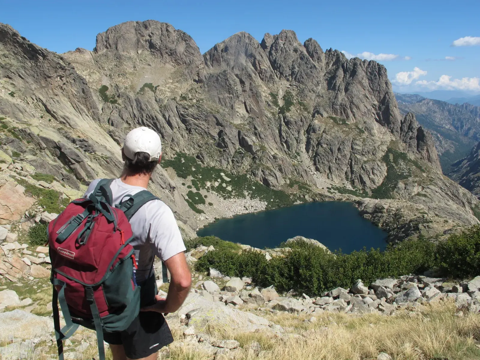 Wanderer mit Rucksack blickt während einer Trekkingtour auf dem GR20 auf Korsika auf einen tiefblauen Bergsee in schroffer Felslandschaft.