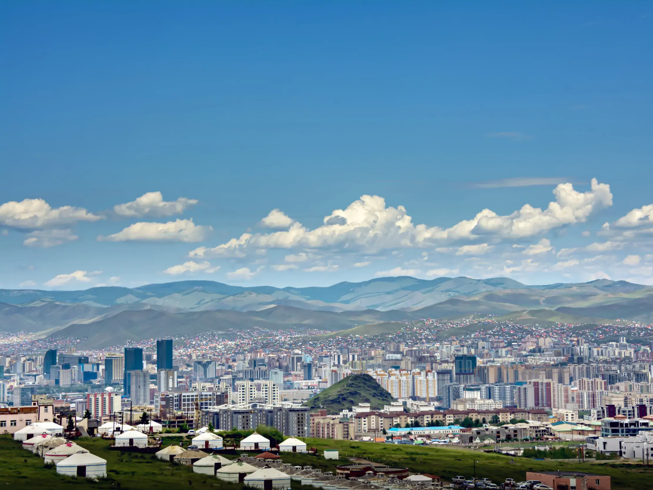 Traditionelle Jurten auf einem grünen Hügel mit Blick auf die moderne Skyline der Metropole Ulaanbaatar in der Mongolei.