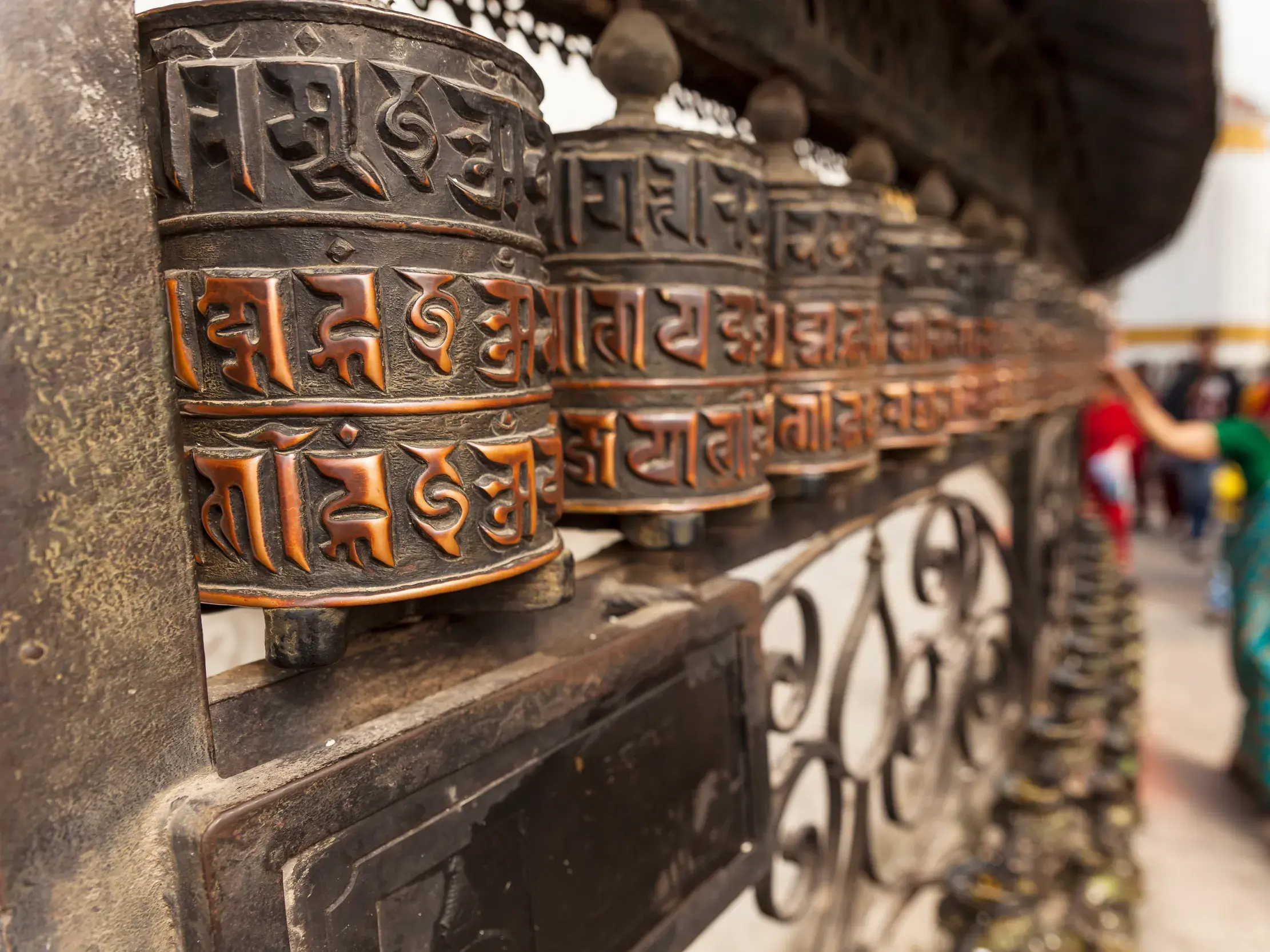 Nahaufnahme von metallenen Gebetsmühlen am Swayambhunath-Stupa in Kathmandu. Eine Frau dreht sie im Vorbeigehen.