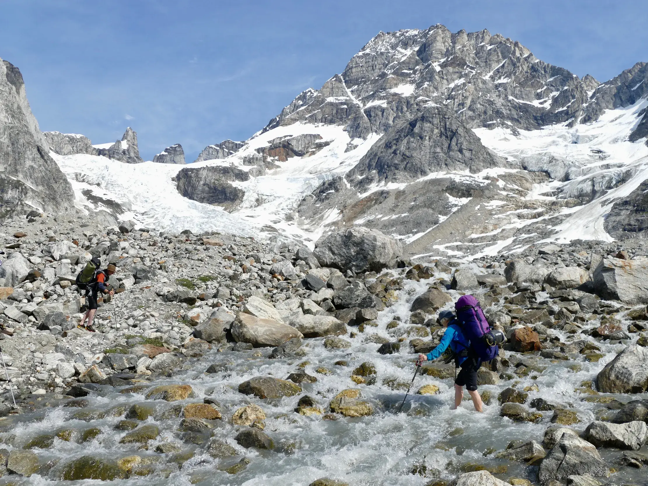 Zwei Trekker durchqueren eine felsige Gletscherlandschaft in Ostgrönland, eine Person watet durch einen kalten Gebirgsbach.