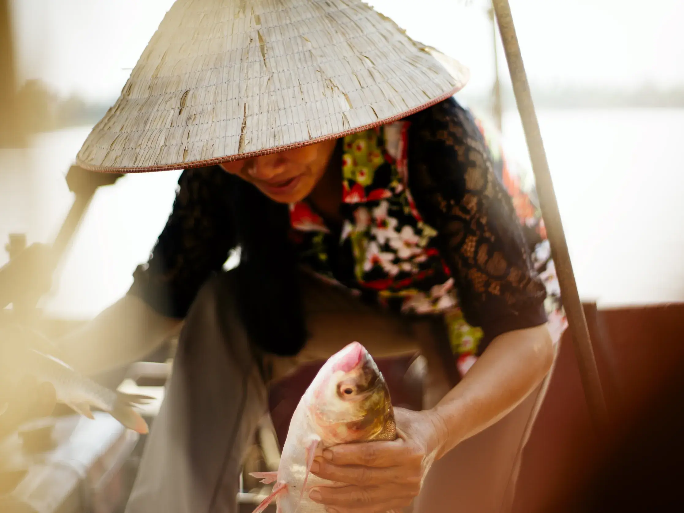 Vietnamesin mit traditionellem Kegelhut hält auf einem Boot im Mekong einen frisch gefangenen Fisch in den Händen.