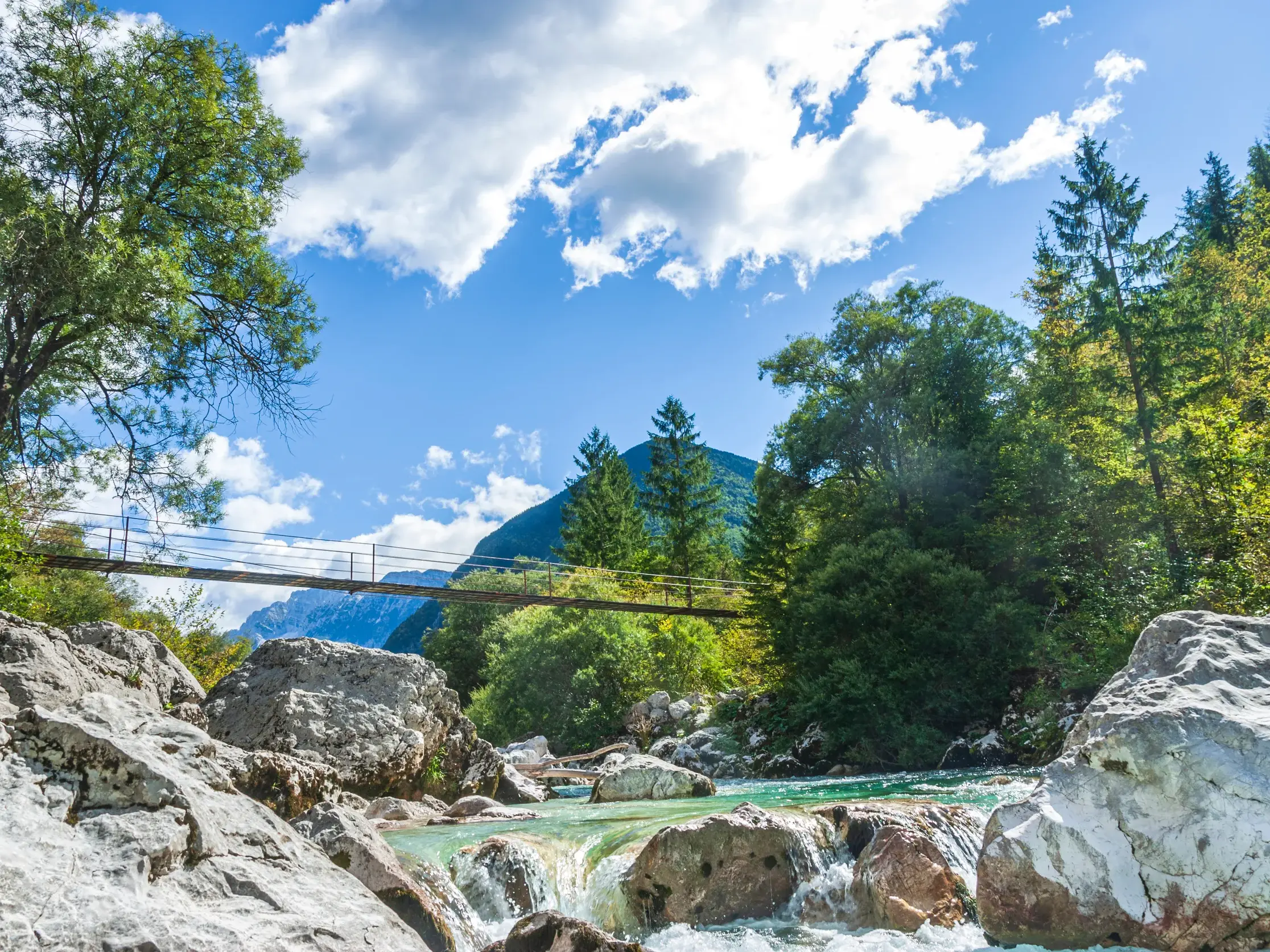 Sonnige Flusslandschaft im Soča-Tal mit einer Hängebrücke über die türkisfarbene Soča und den Julischen Alpen im Hintergrund.