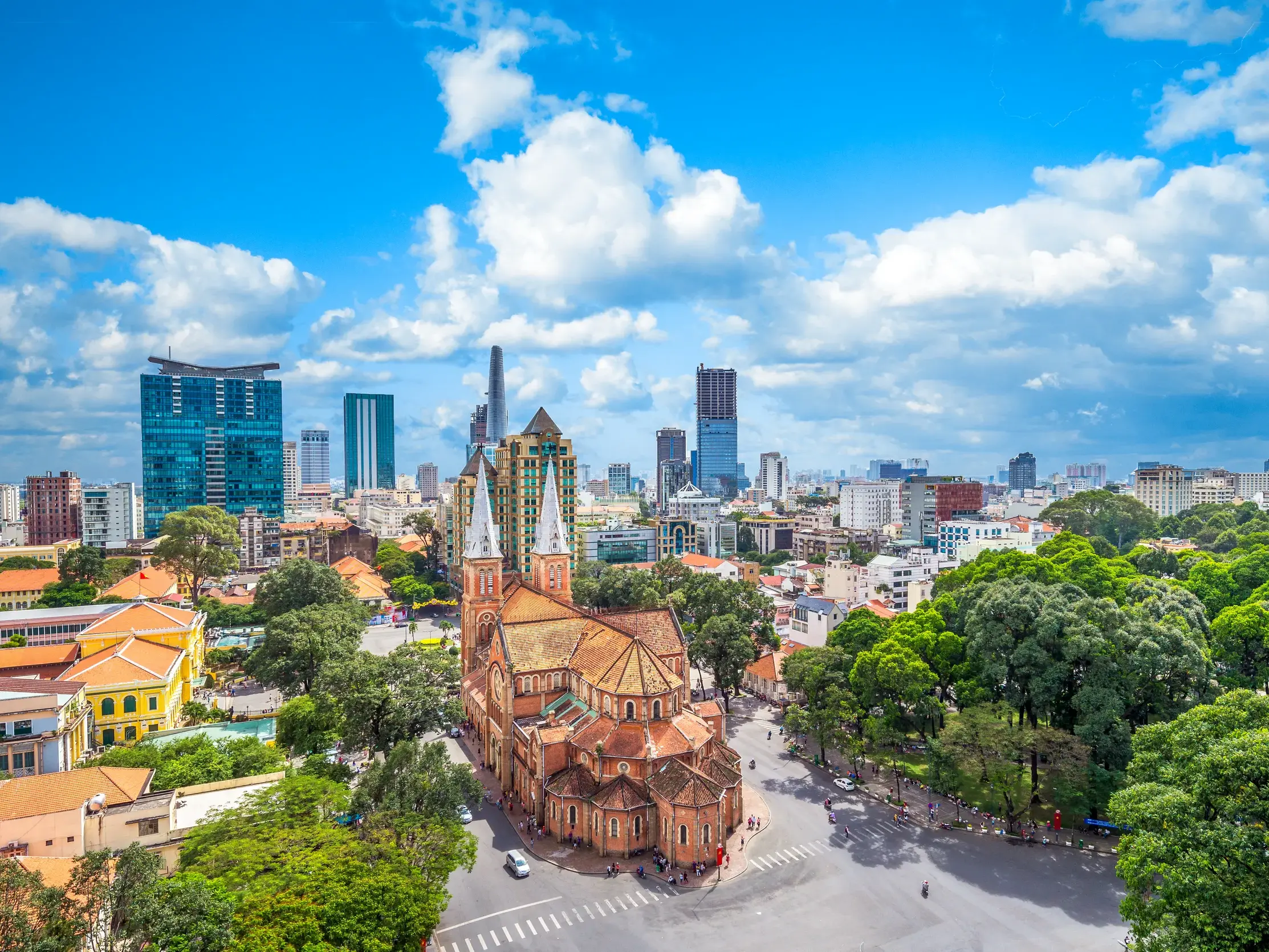 Die historische Kathedrale Notre-Dame in Saigon vor der modernen Skyline mit Wolkenkratzern unter blauem Himmel.