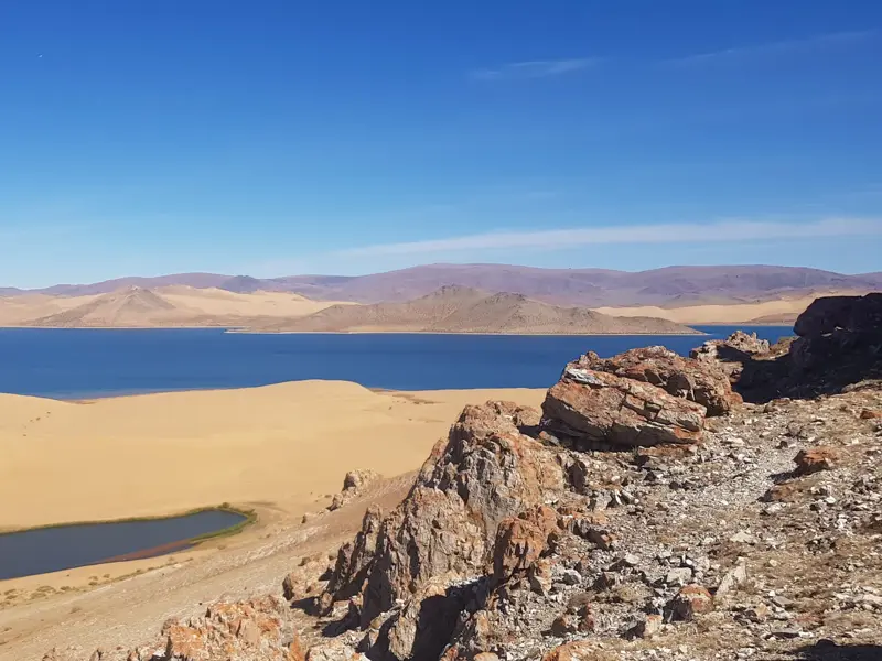 Blick von einer Felsklippe auf den tiefblauen Khar-See in der Mongolei, umgeben von Sanddünen und sanften Bergen am Horizont.