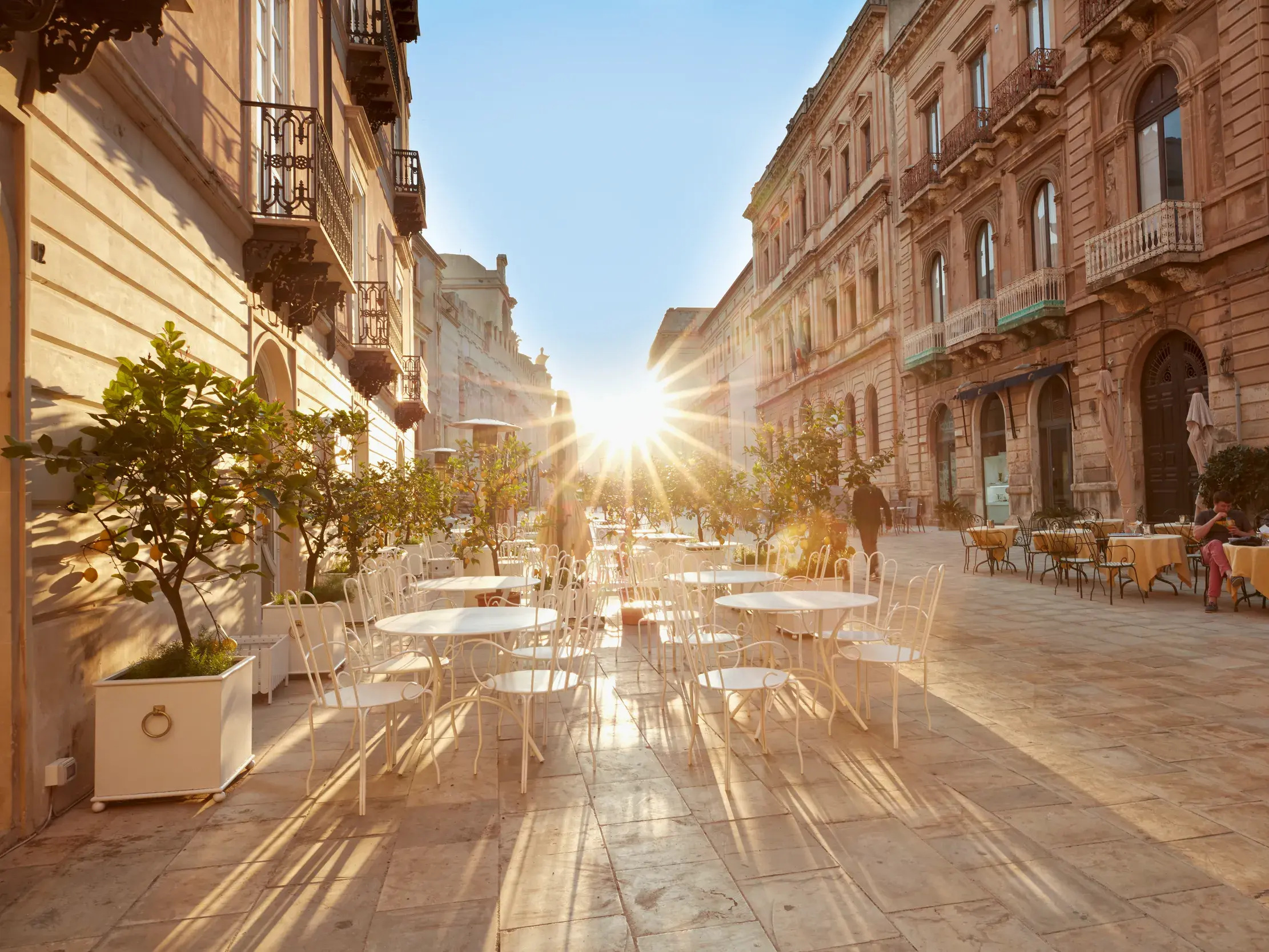Ein Straßencafé in der Altstadt von Syrakus auf Ortygia, dessen weiße Tische von der tiefstehenden Sonne angestrahlt werden.