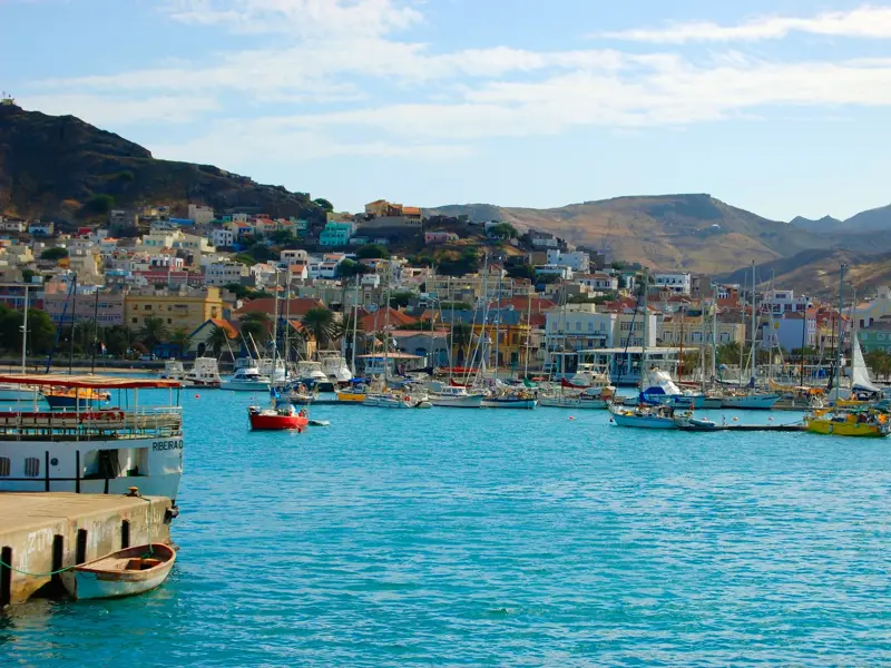 Zahlreiche Boote im türkisblauen Wasser des Hafens von Mindelo auf São Vicente, im Hintergrund die bunten Häuser der Stadt am Hang.