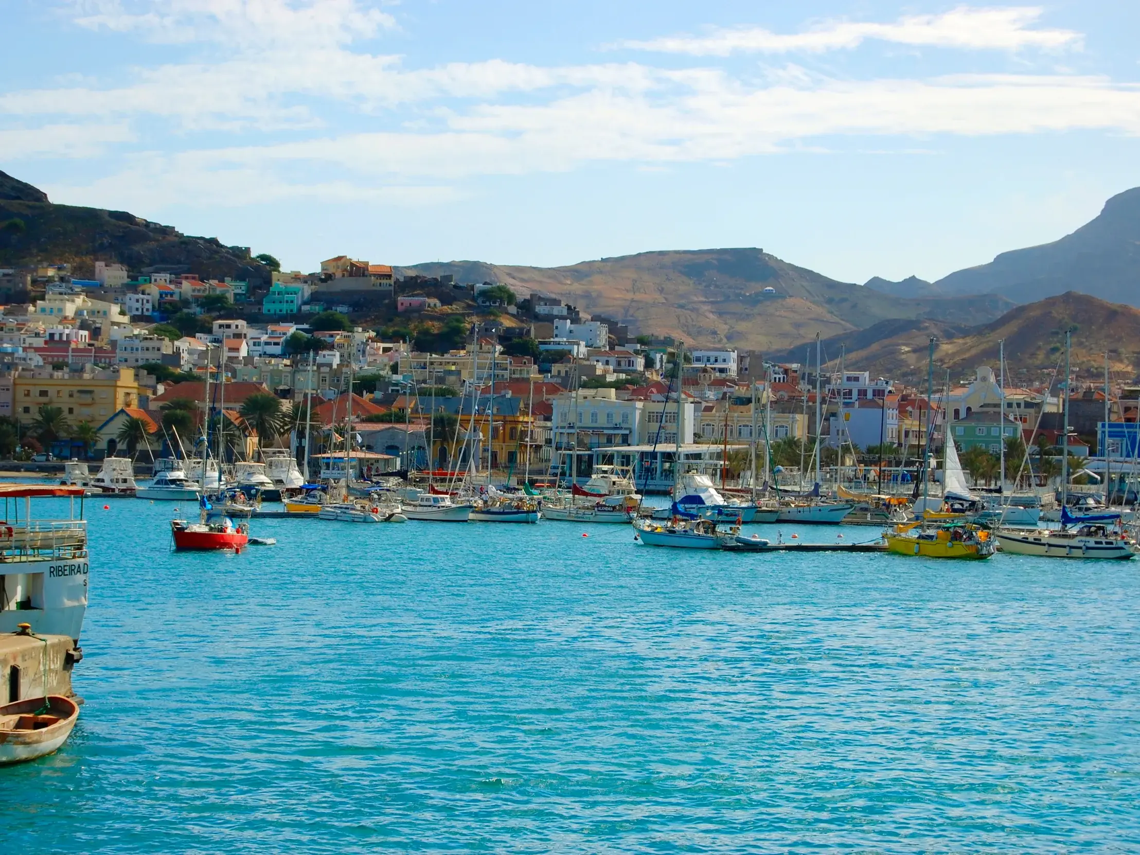 Hafen von Mindelo auf São Vicente mit Segelbooten im türkisen Wasser. Im Hintergrund die am Hang gebaute bunte Stadt.