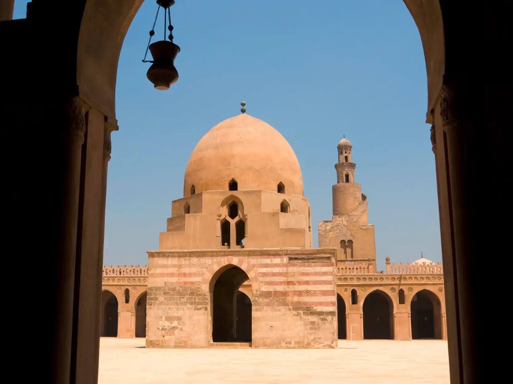 Blick durch einen Torbogen auf den sonnigen Hof der Ibn-Tulun-Moschee in Kairo mit dem Kuppelbau und dem Minarett.