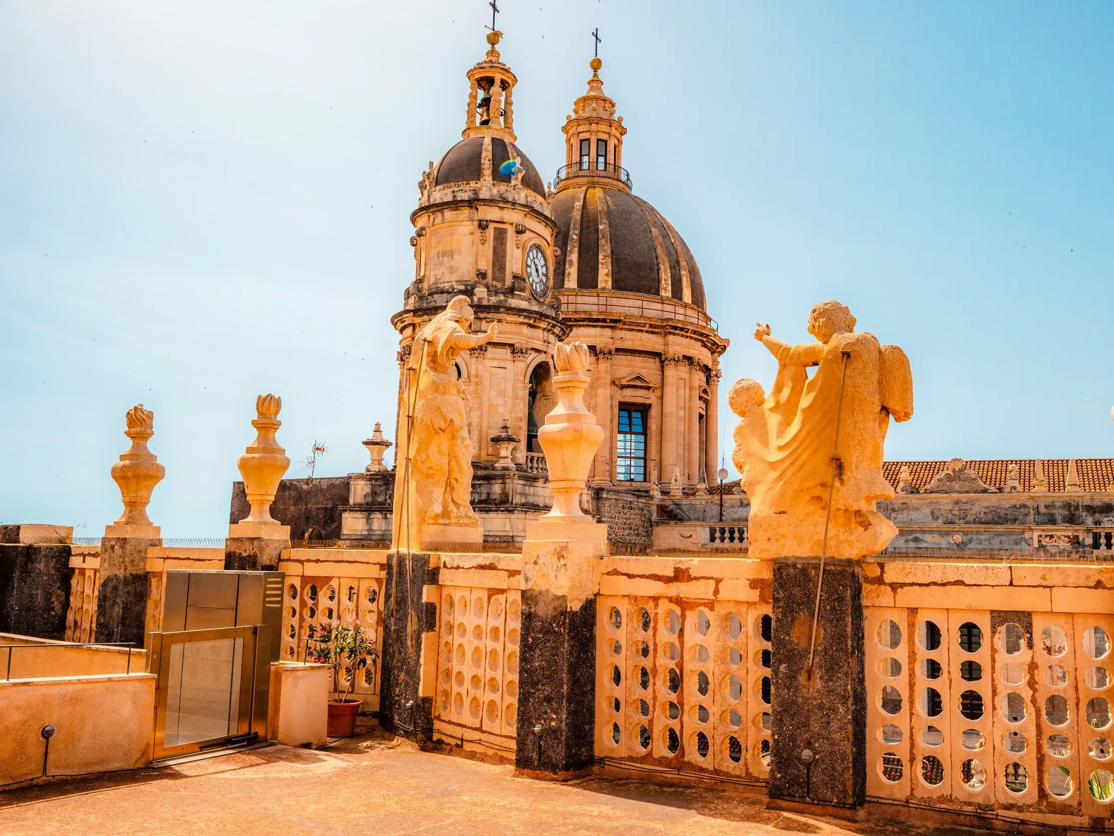 Sonniger Ausblick von einer Terrasse mit Steinskulpturen auf die prachtvollen Barockkuppeln der Kathedrale von Catania.