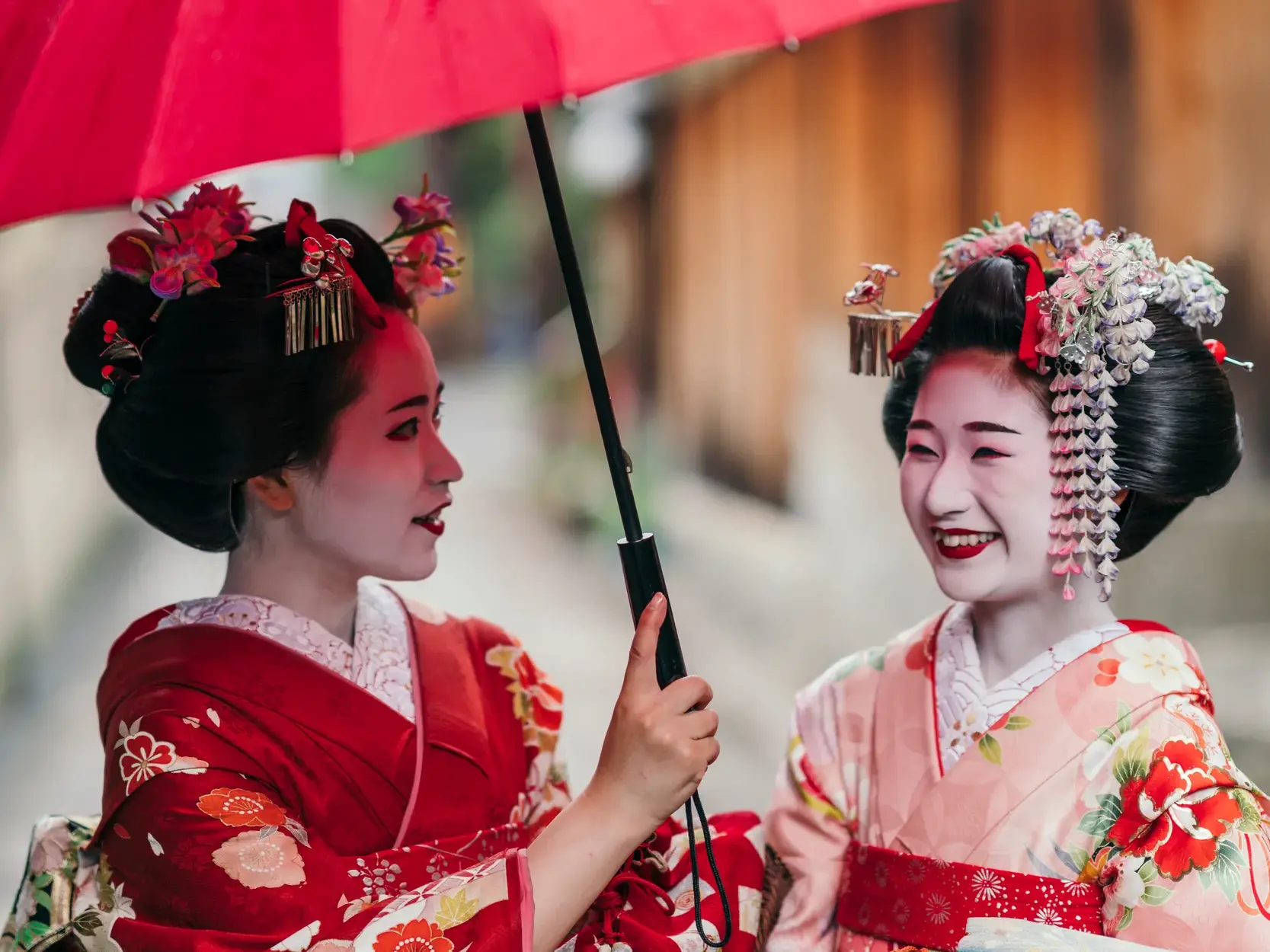 Zwei Geishas in traditionellen Kimonos unterhalten sich lächelnd unter einem roten Schirm im Geisha-Viertel Gion in Kyoto.