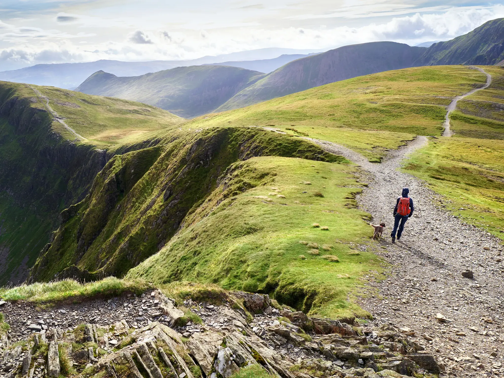 Ein Wanderer mit Hund auf einem Bergpfad, der über die weiten, grünen Hügel des Lake District in England führt.