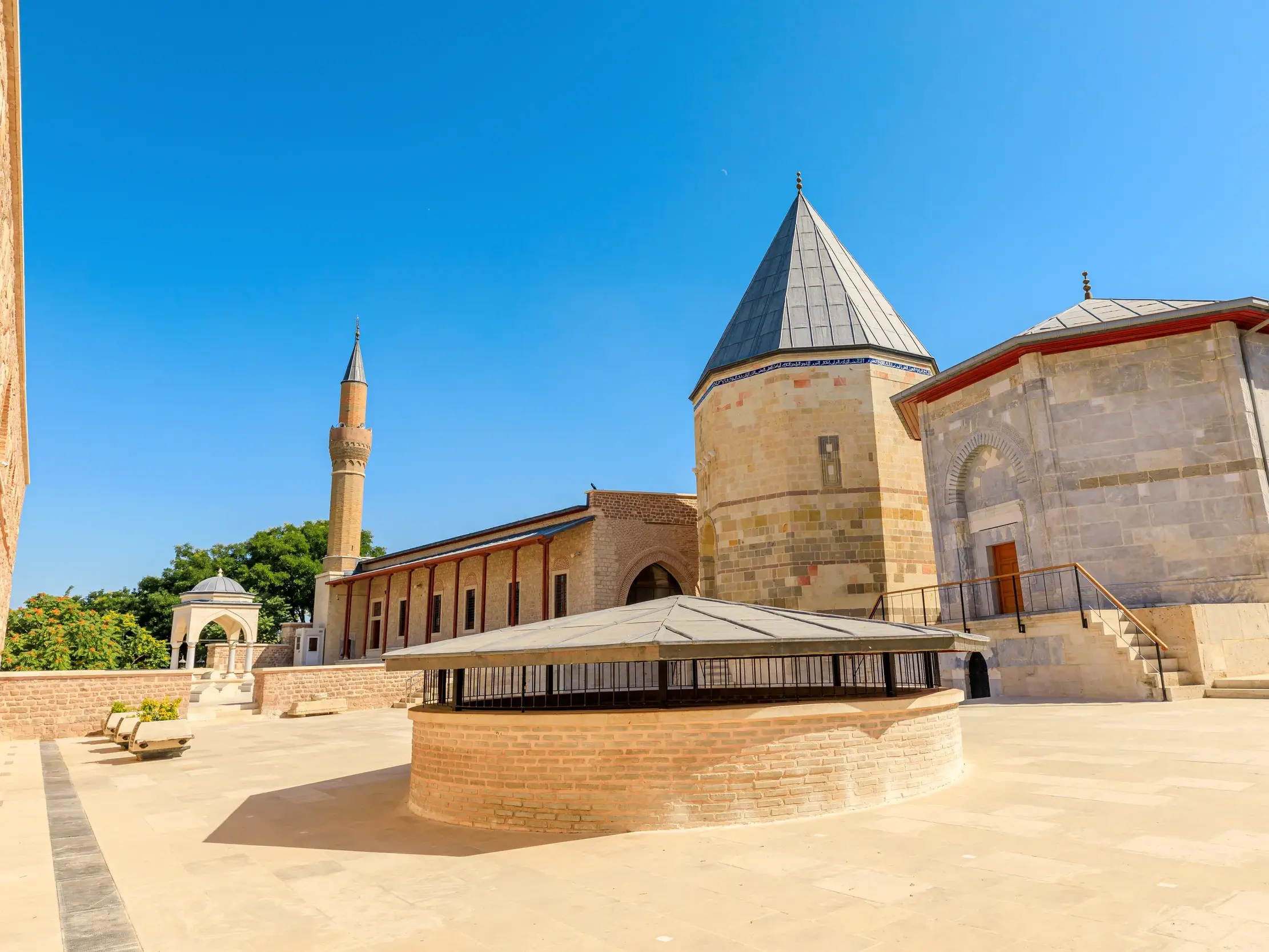 Sonniger Innenhof des Mevlana-Museums in Konya mit dem Mausoleum des Mystikers Rumi und einem Minarett unter blauem Himmel.