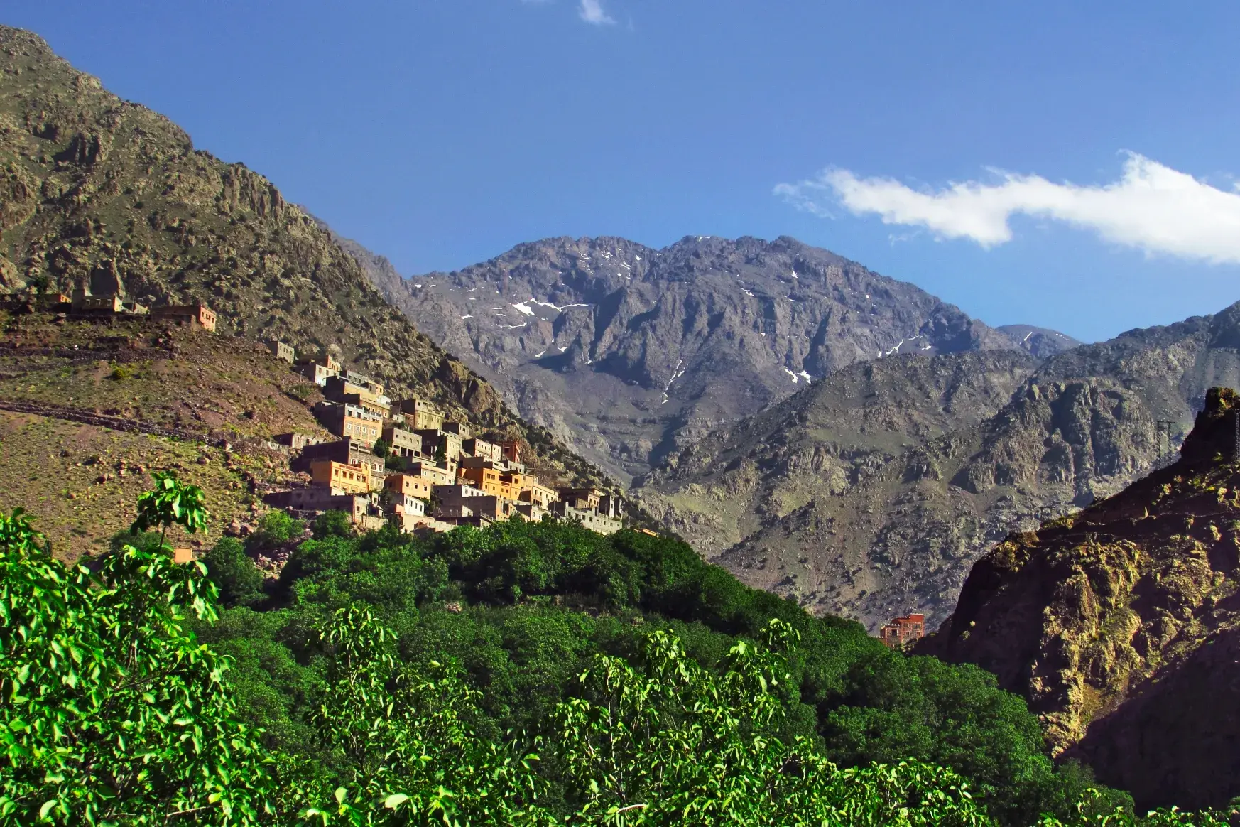 Sonnenbeschienenes Berberdorf an einem Berghang im Hohen Atlas vor der Kulisse des schneebedeckten Djebel Toubkal.