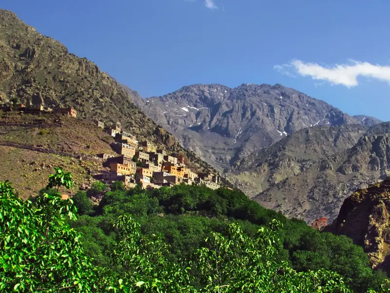 Ein Berberdorf schmiegt sich an einen Hang im Hohen Atlas, im Hintergrund der schneebedeckte Gipfel des Djebel Toubkal.