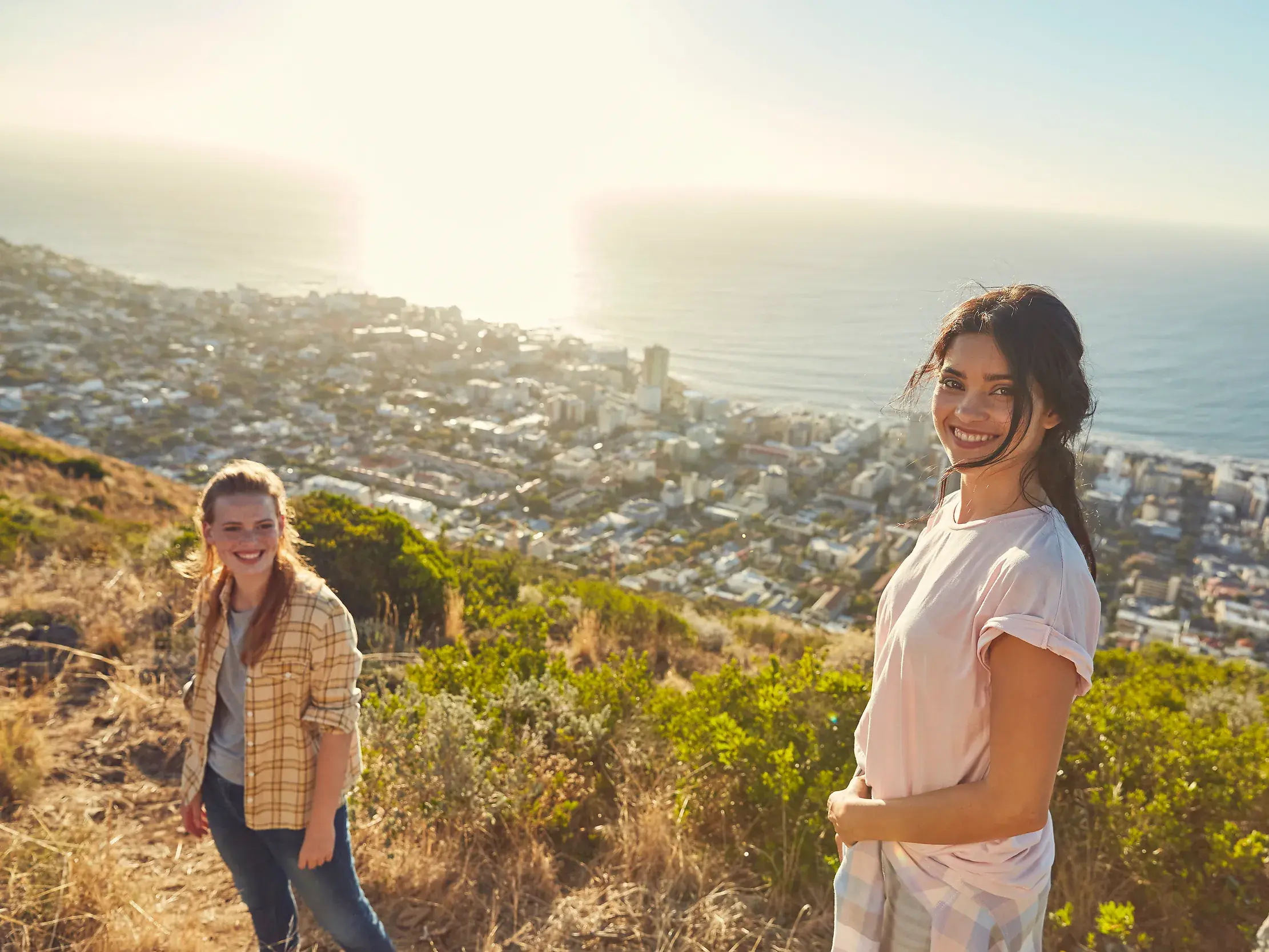 Zwei junge Frauen einer Gruppenreise genießen auf dem Signal Hill den sonnigen Panoramablick über Kapstadt und das Meer.